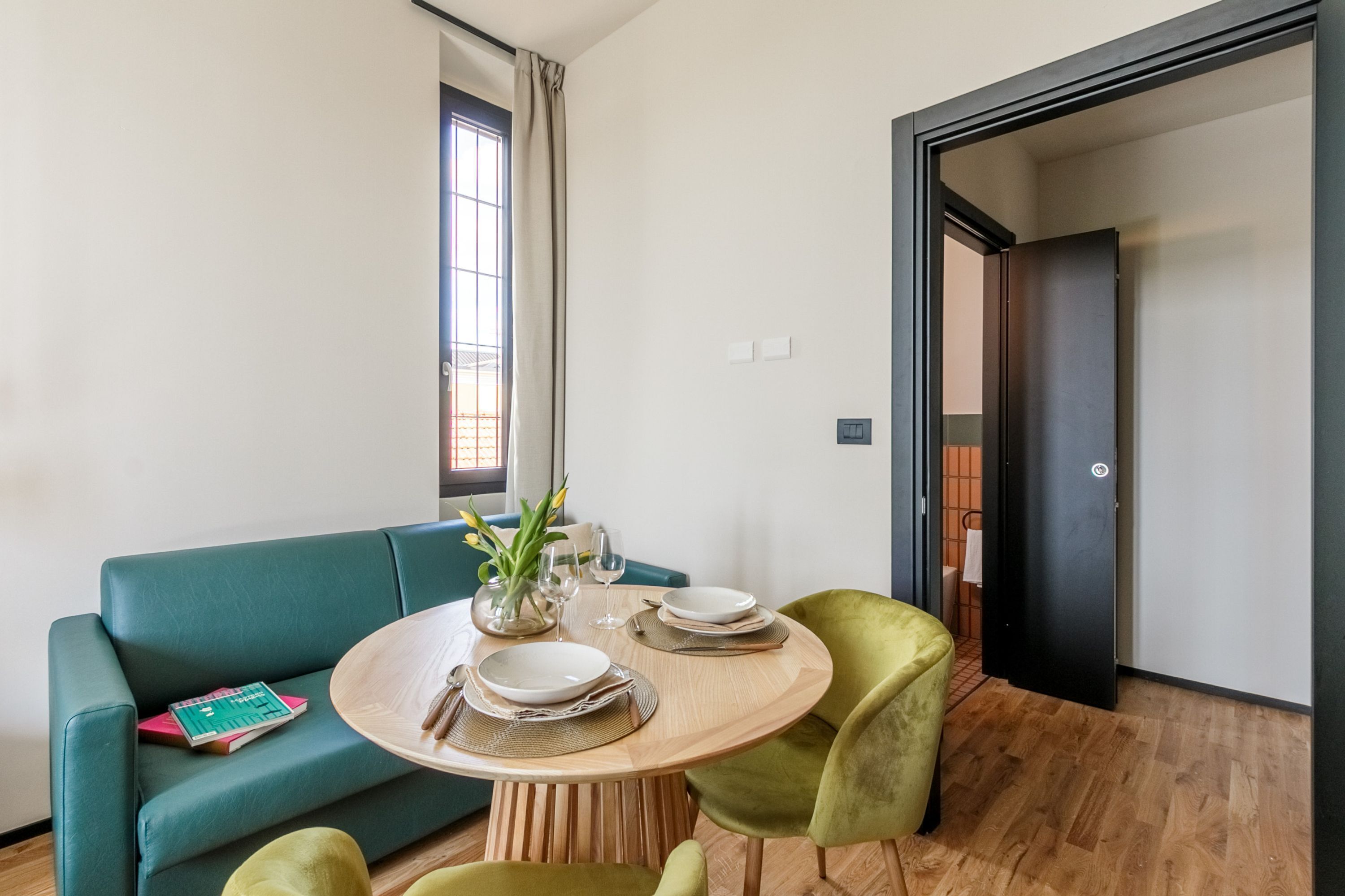 A modern room with a round wooden dining table, green upholstered sofa, and a small side table with a vase and books.