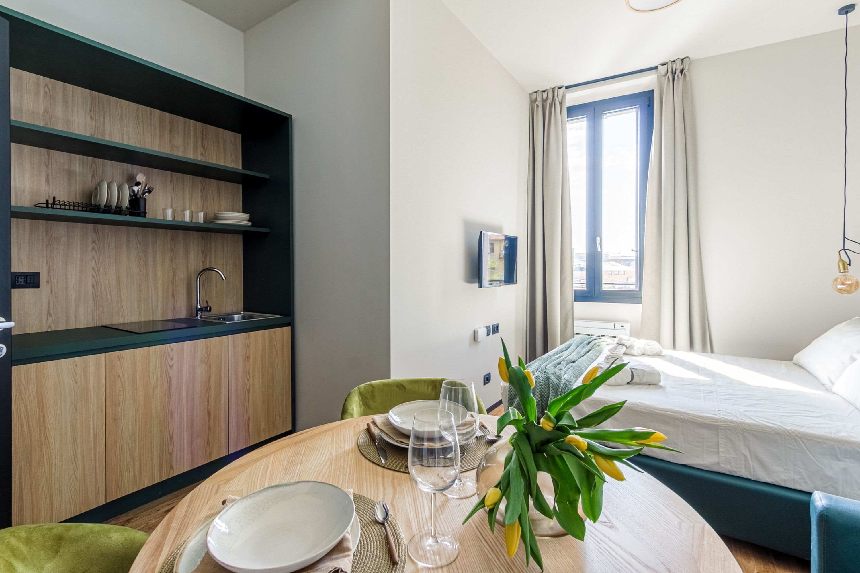 Quarto de hotel moderno com uma mesa de jantar de madeira posta para dois, uma cama com cabeceira azul e uma janela com cortinas.