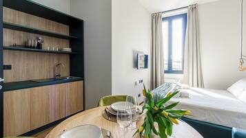 Quarto de hotel moderno com uma mesa de jantar de madeira posta para dois, uma cama com cabeceira azul e uma janela com cortinas.