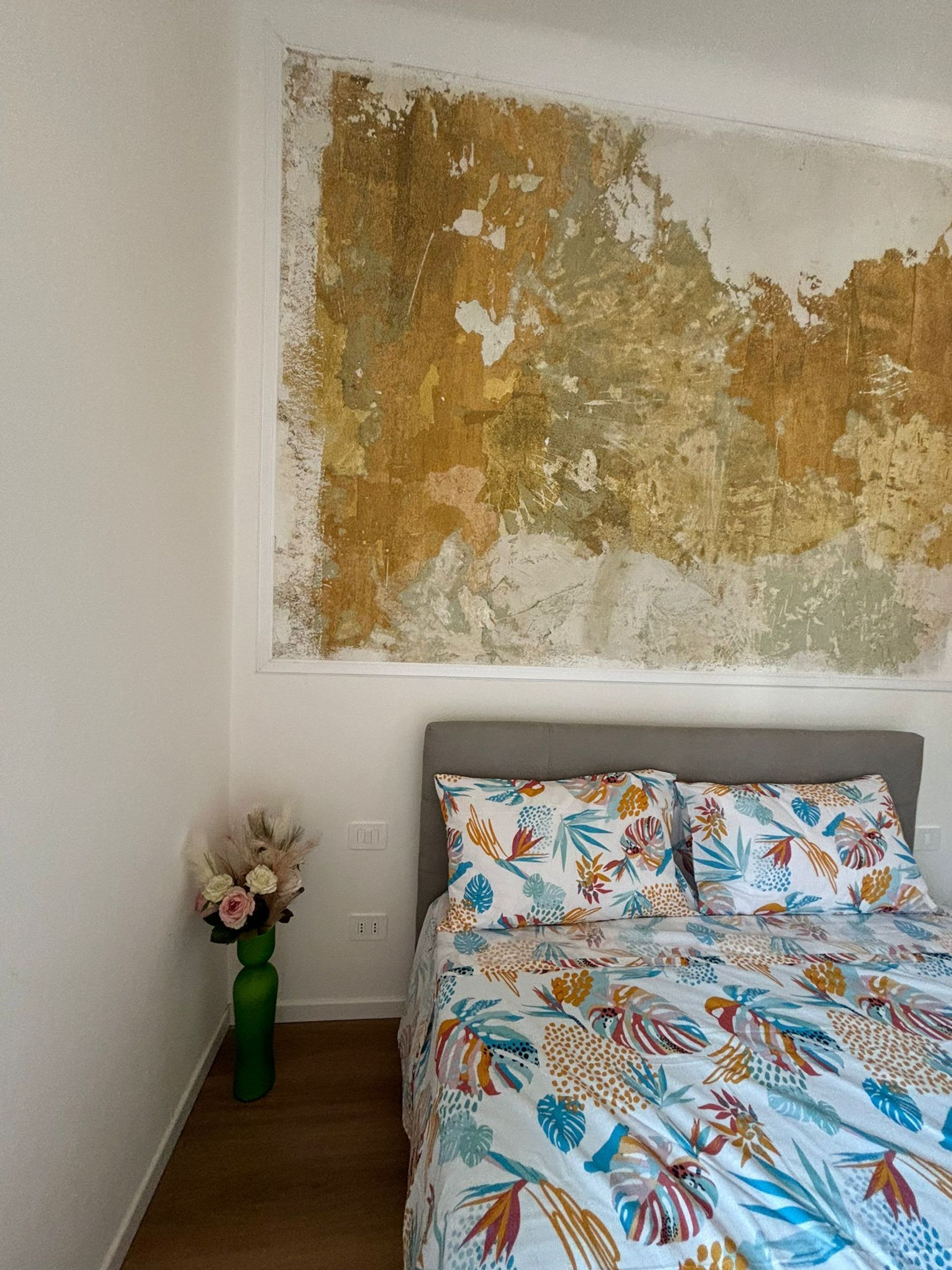 A bedroom with a bed, a green vase with flowers, and a large abstract painting on the wall.