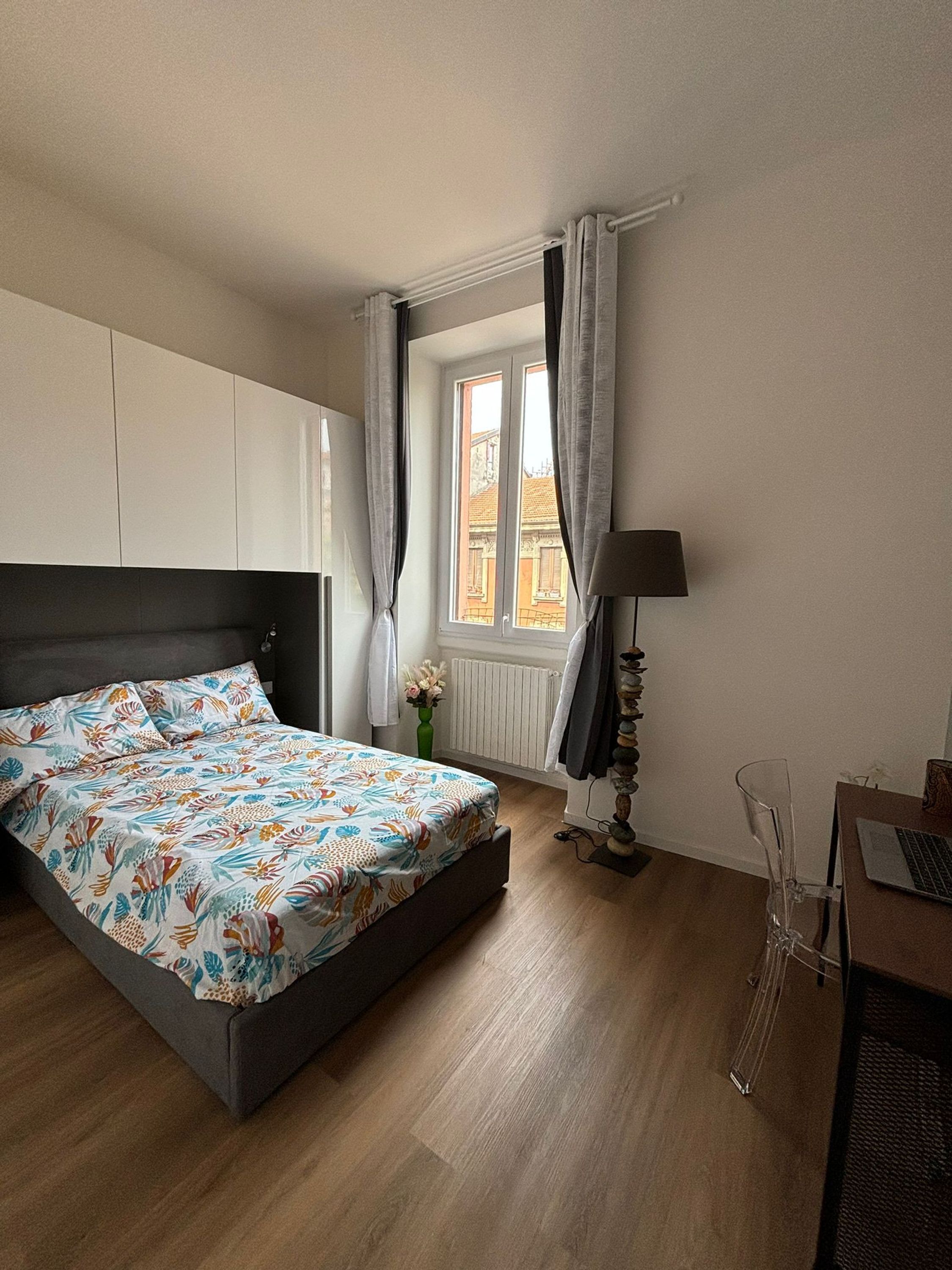 A bedroom with a bed, a desk with a laptop, a lamp, and a window with curtains.