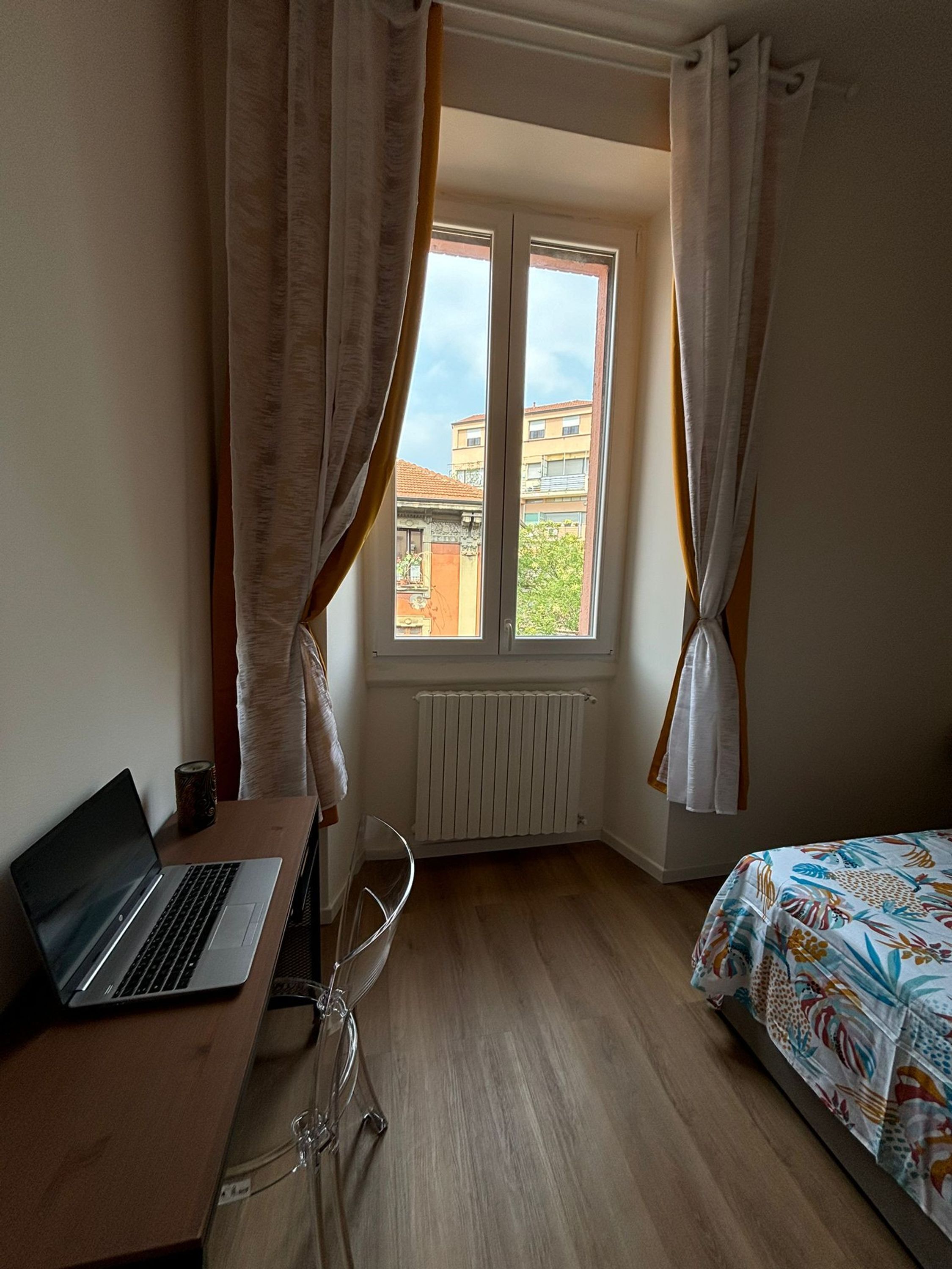 Ein Schlafzimmer mit einem Bett, einem Schreibtisch mit einem Laptop, einem Heizkörper und einem Fenster mit Vorhängen.