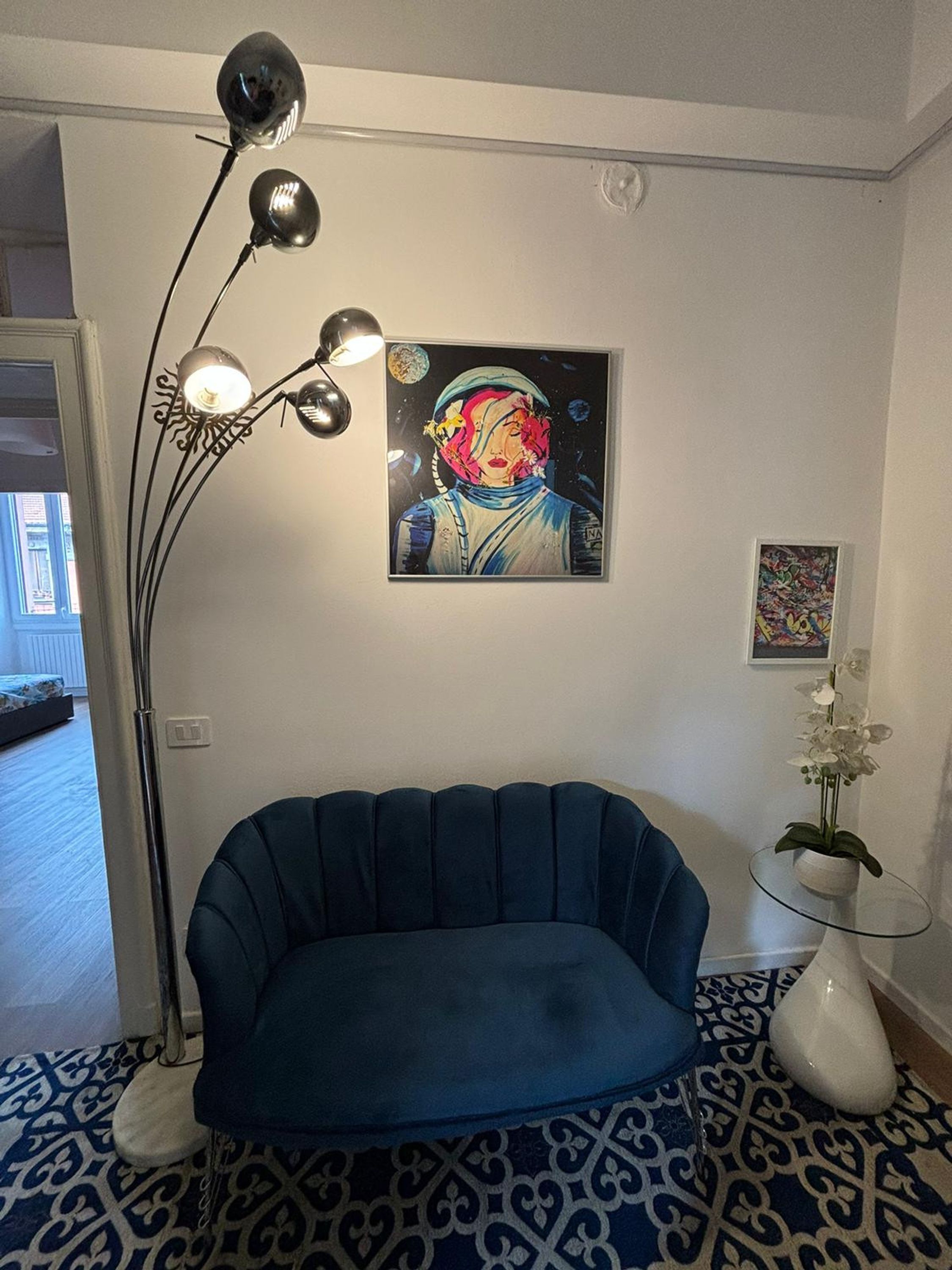 Ein Zimmer mit einem blau gepolsterten Sofa, einer Stehlampe, einem Gemälde einer Frau in einem Raumanzug und einer kleinen Orchideenpflanze auf einem Glastisch.