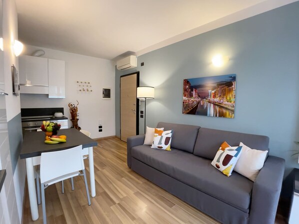 A modern living room with a grey sofa, a dining table with fruit, and a cityscape painting on the wall. - Candiani F1 Bilocale (Milan)