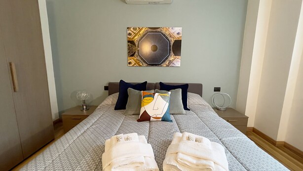 A hotel room with a bed, a wall-mounted air conditioner, a decorative wall piece, a bedside table with a lamp, and a round mirror. - Candiani F1 Bilocale (Milan)