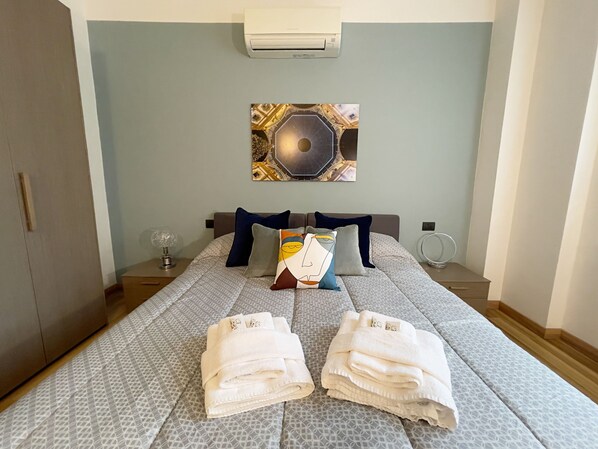 A hotel room with a bed, a wall-mounted air conditioner, a decorative wall piece, a bedside table with a lamp, and a round mirror. - Candiani F1 Bilocale (Milan)