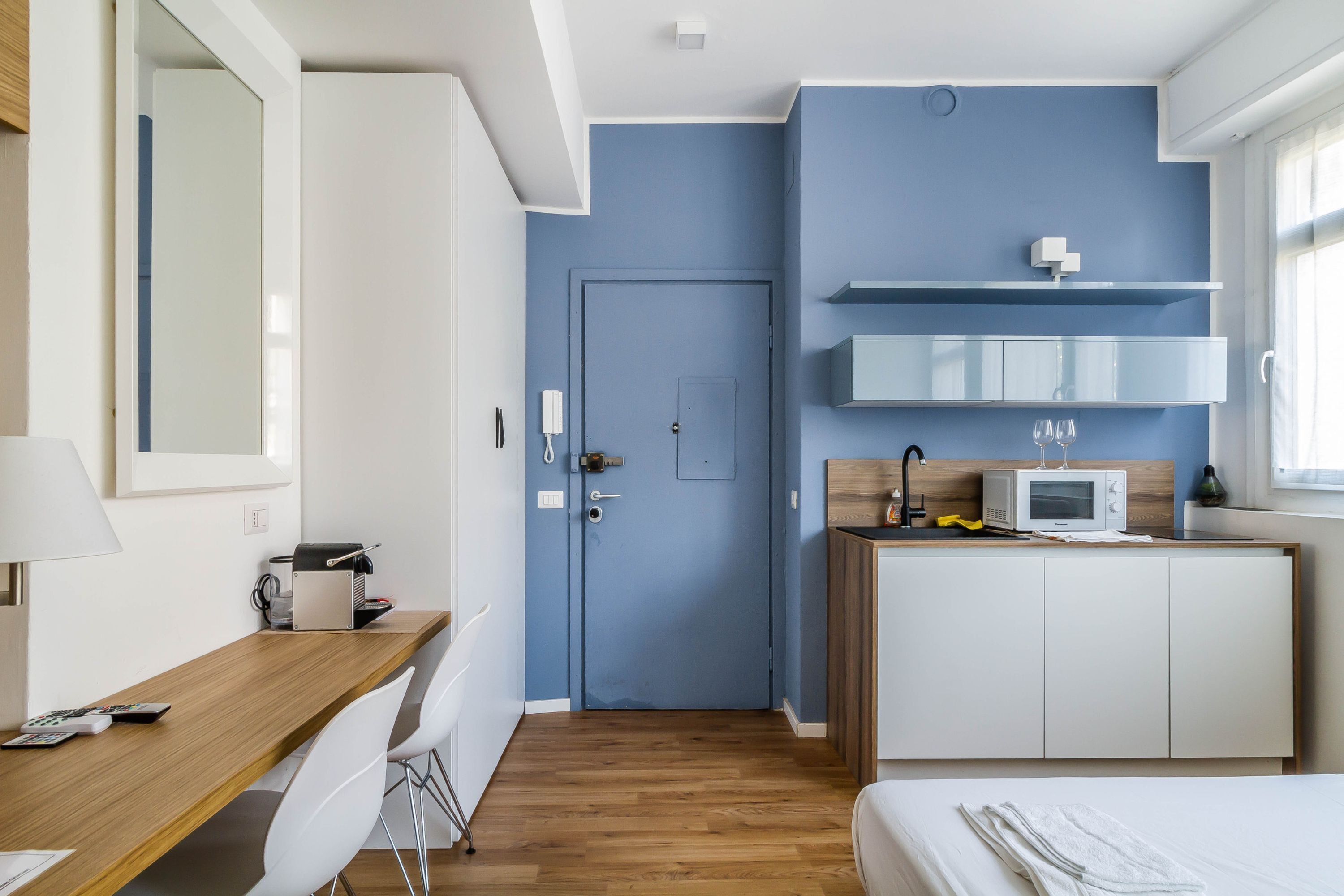 Habitación de hotel compacta con escritorio de madera, sillas blancas, cama, microondas y una pequeña cocina americana.