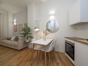 A modern living room with a white sofa, a dining table with chairs, a potted plant, a floor lamp, and a kitchen area with an oven. - Porta Vittoria - Cozy Stylish 1bdr in Duomo San Babila Area (Milan)