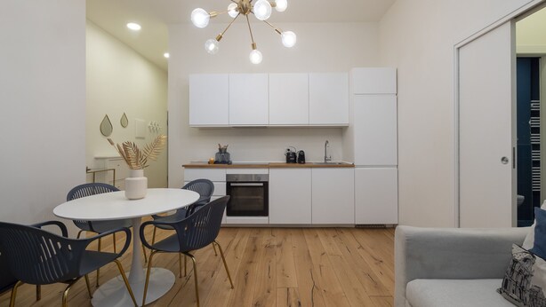 A modern kitchen with white cabinets, a built-in oven, and a sink. There's a dining area with a round table and chairs, and a grey sofa in the living area. - Porta Vittoria - Cozy Stylish 1bdr in Duomo San Babila Area (Milan)