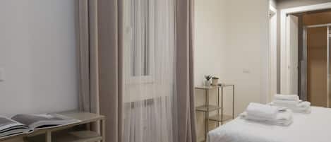 A hotel room with a bed, a bedside table, a small table with a plant, and a window with sheer curtains.