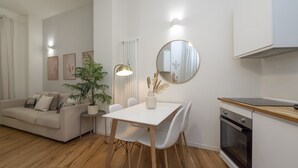 Una sala moderna con un sofá blanco, una mesa de comedor con sillas, una planta en maceta, una lámpara de pie y una zona de cocina con horno.