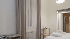 Una habitación de hotel con cama, una mesita de noche, una pequeña mesa con una planta y una ventana con cortinas traslúcidas.