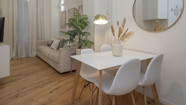 Una sala moderna con un sofá blanco, una mesa de comedor con sillas, una planta en maceta, una lámpara de pie y un espejo redondo en la pared.