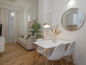 Ein modernes Wohnzimmer mit einem weißen Sofa, einem Esstisch mit Stühlen, einer Topfpflanze, einer Stehlampe und einem runden Spiegel an der Wand.