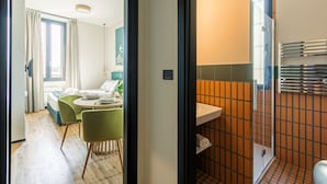 Quarto de hotel moderno com área de refeições, banheiro com chuveiro revestido em azulejos e vista para o exterior.