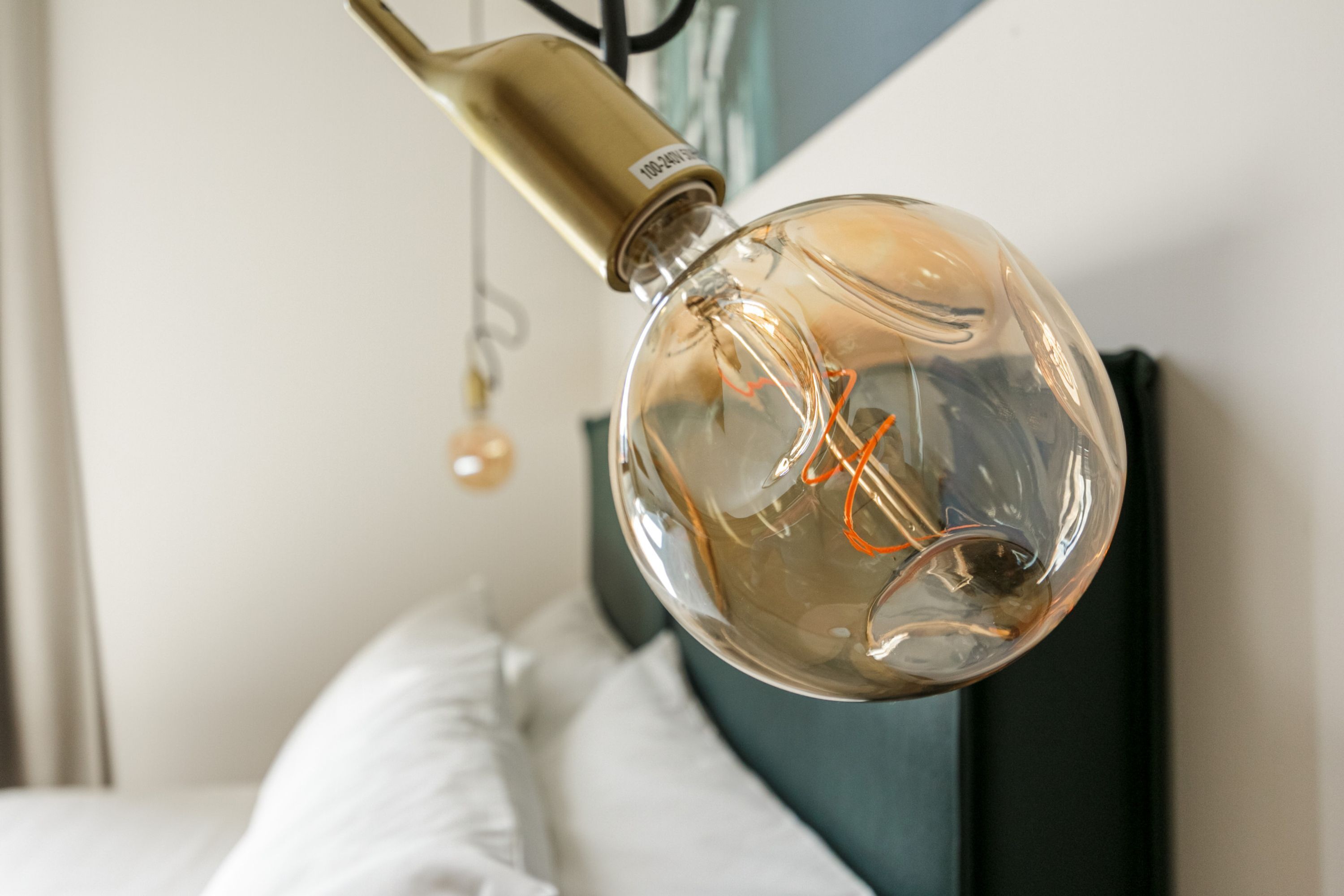 Une ampoule transparente, avec un filament visible et une base en laiton, suspendue à un cordon noir dans une pièce où se trouvent un lit et une tête de lit sombre.