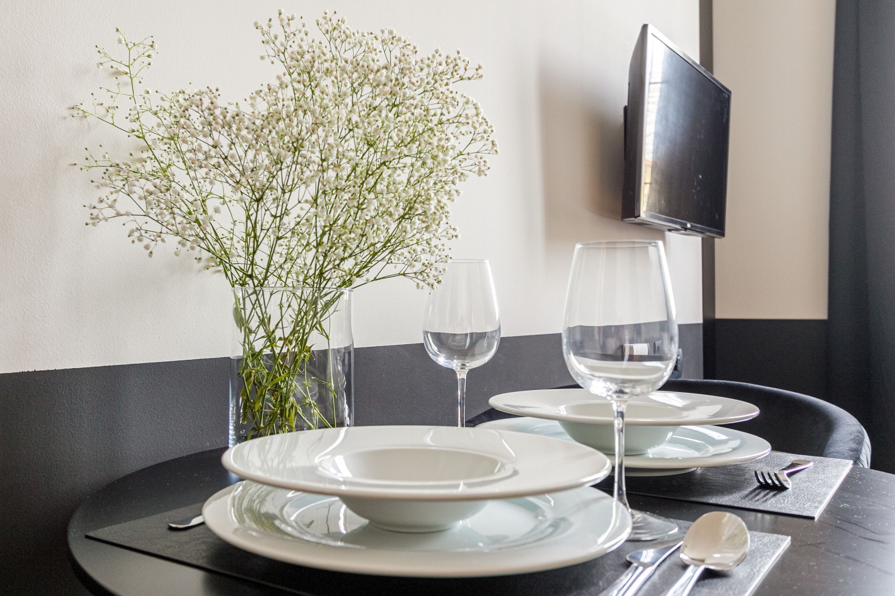 A dining table set with plates, glasses, and cutlery, a vase with white flowers, and a flat-screen TV mounted on the wall.