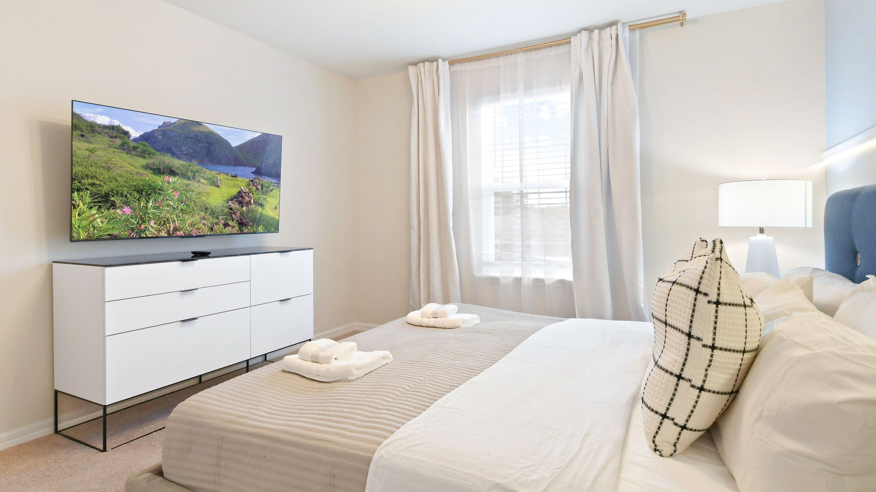 A hotel room with a bed, a TV on a white dresser, a window with curtains, and a lamp.