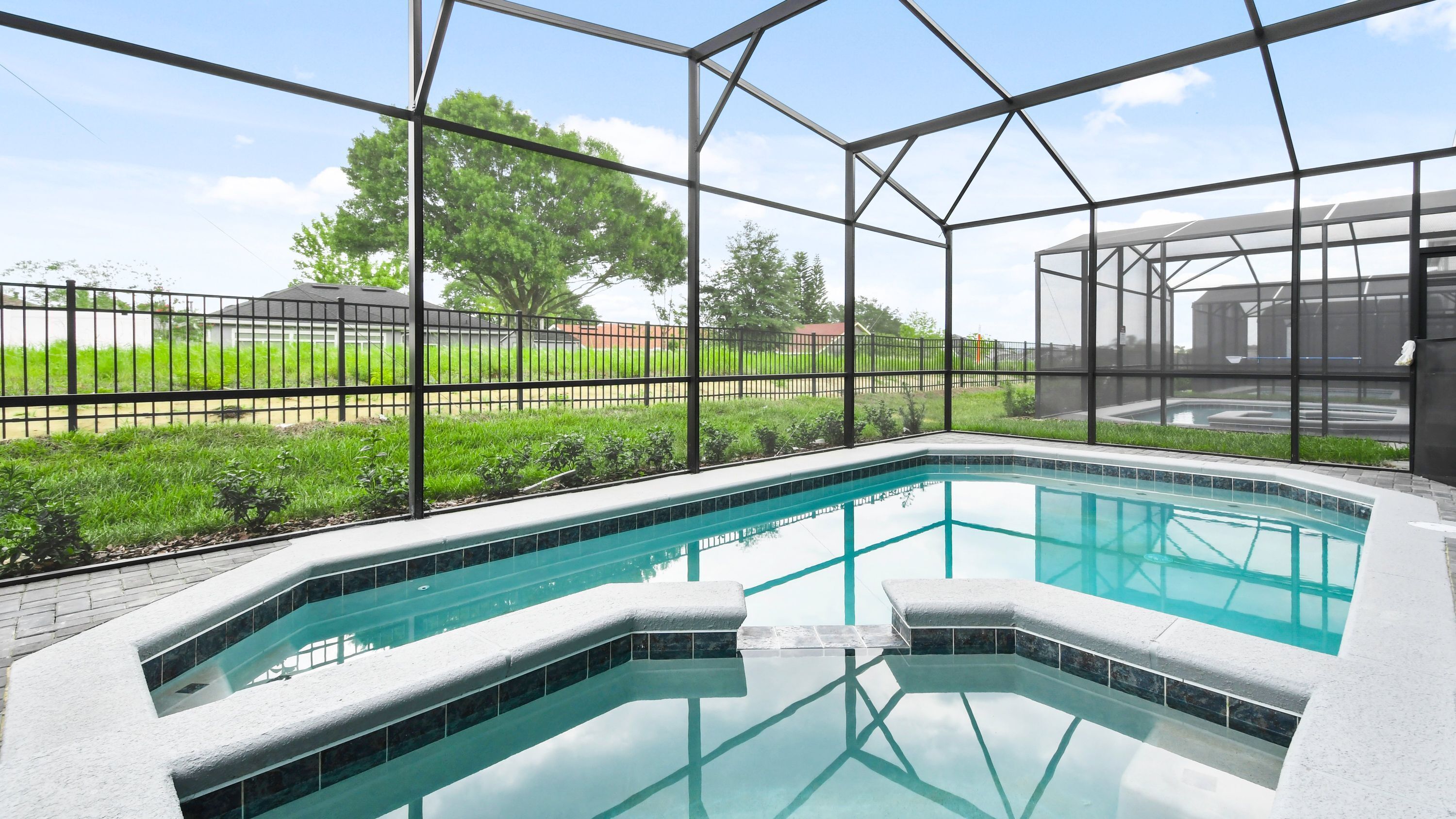 An indoor swimming pool with clear blue water, surrounded by a metal frame structure and a view of a green landscape with trees and a fence.