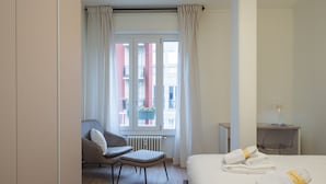 Habitación de hotel con cama, una silla, una mesita con lámpara y ventana con cortinas.