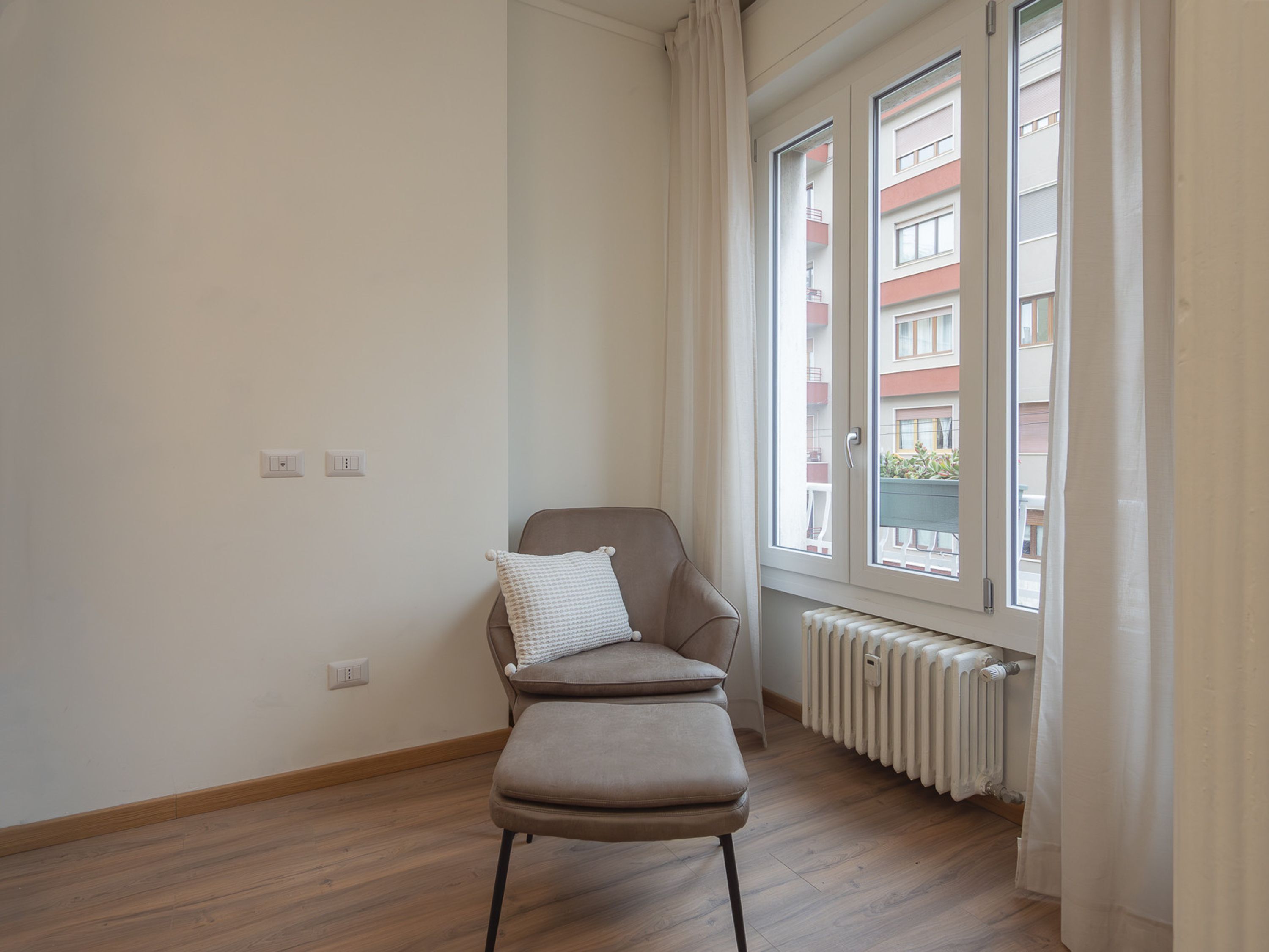 A room with a wooden floor, a single chair with a cushion, a radiator, and a window with curtains.