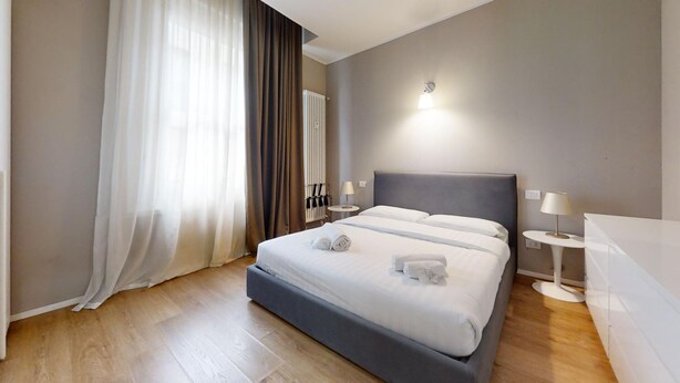 Una habitación de hotel moderna con una cama grande, dos mesitas de noche y una ventana con cortinas traslúcidas.