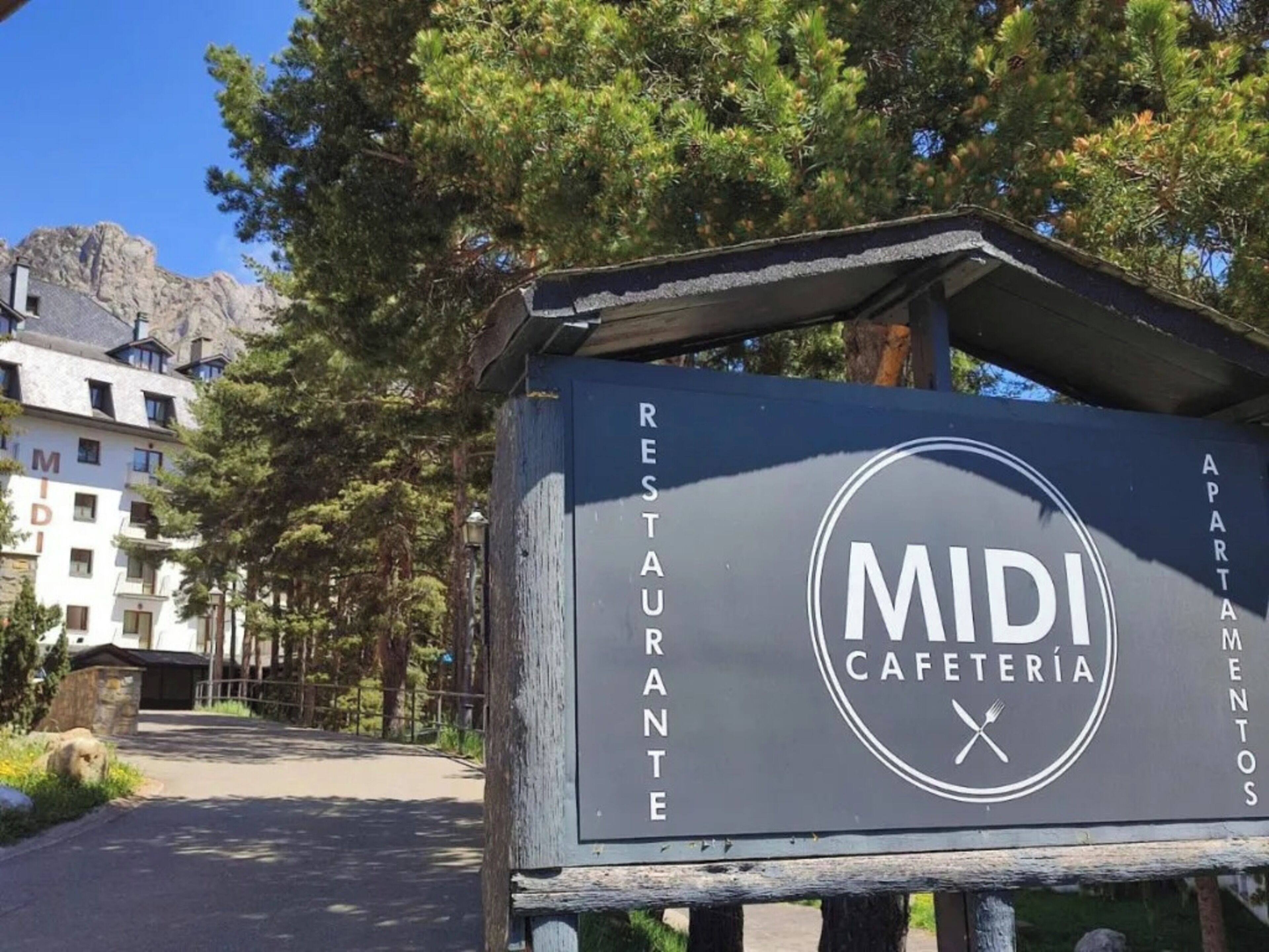 Une enseigne indiquant « MIDI CAFÉTÉRIA », avec une structure en bois rustique, située dans un quartier résidentiel, avec des immeubles d’appartements en arrière-plan.