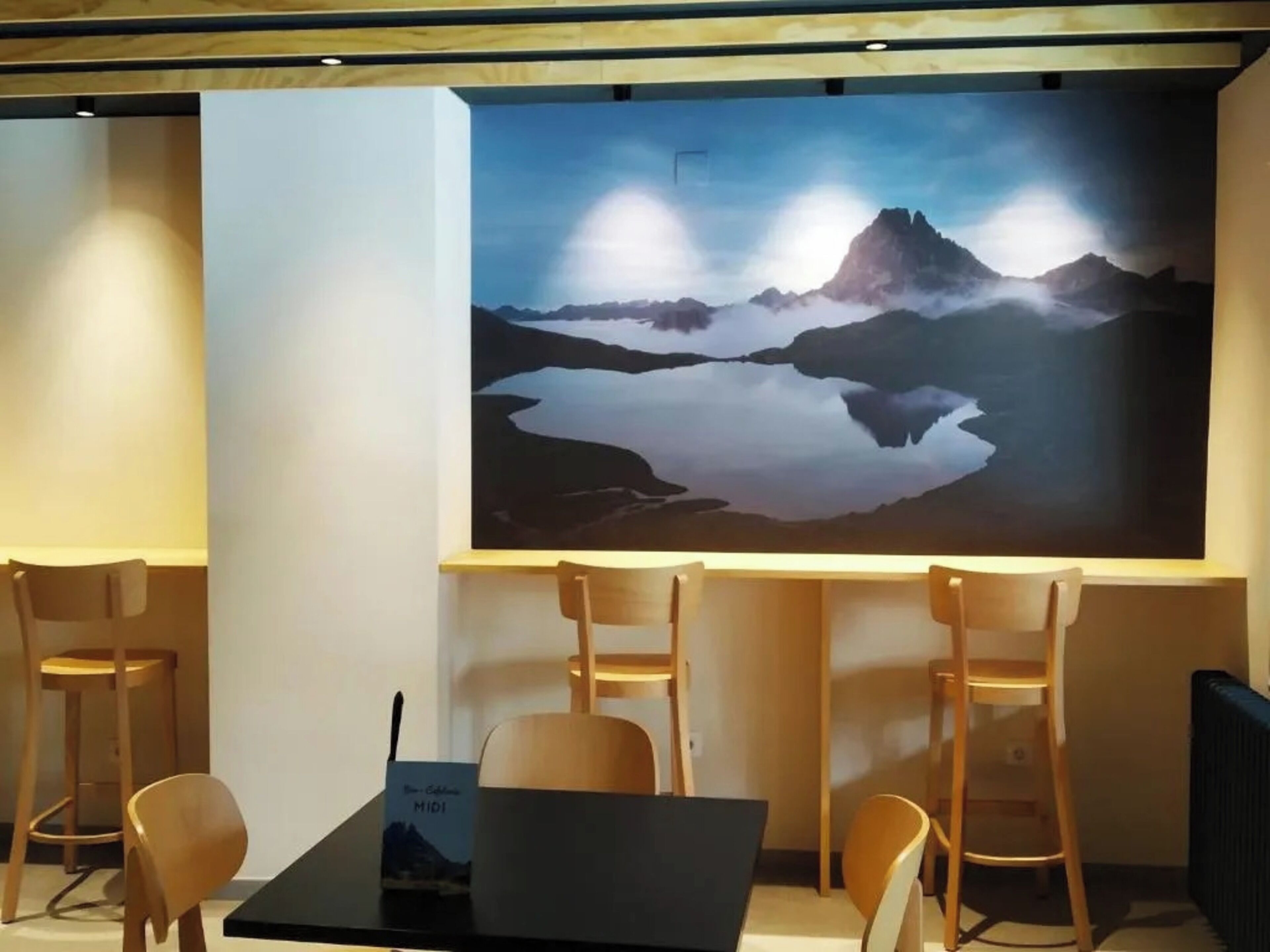 Een moderne kamer met een groot wandmounted scherm waarop een berglandschap te zien is. Er staan houten stoelen en een tafel met een laptop.