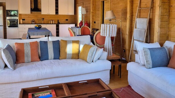 Een gezellig chaletinterieur met een houten plafond, witte banken met opvallende kussens, een keuken met witte kasten en een eettafel met geruit tafelkleed.