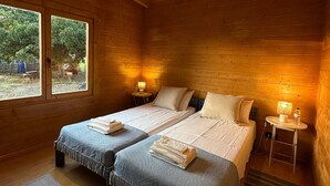 Een houten slaapkamer met twee bedden, een tafeltje met een lamp en een raam met uitzicht op groen.