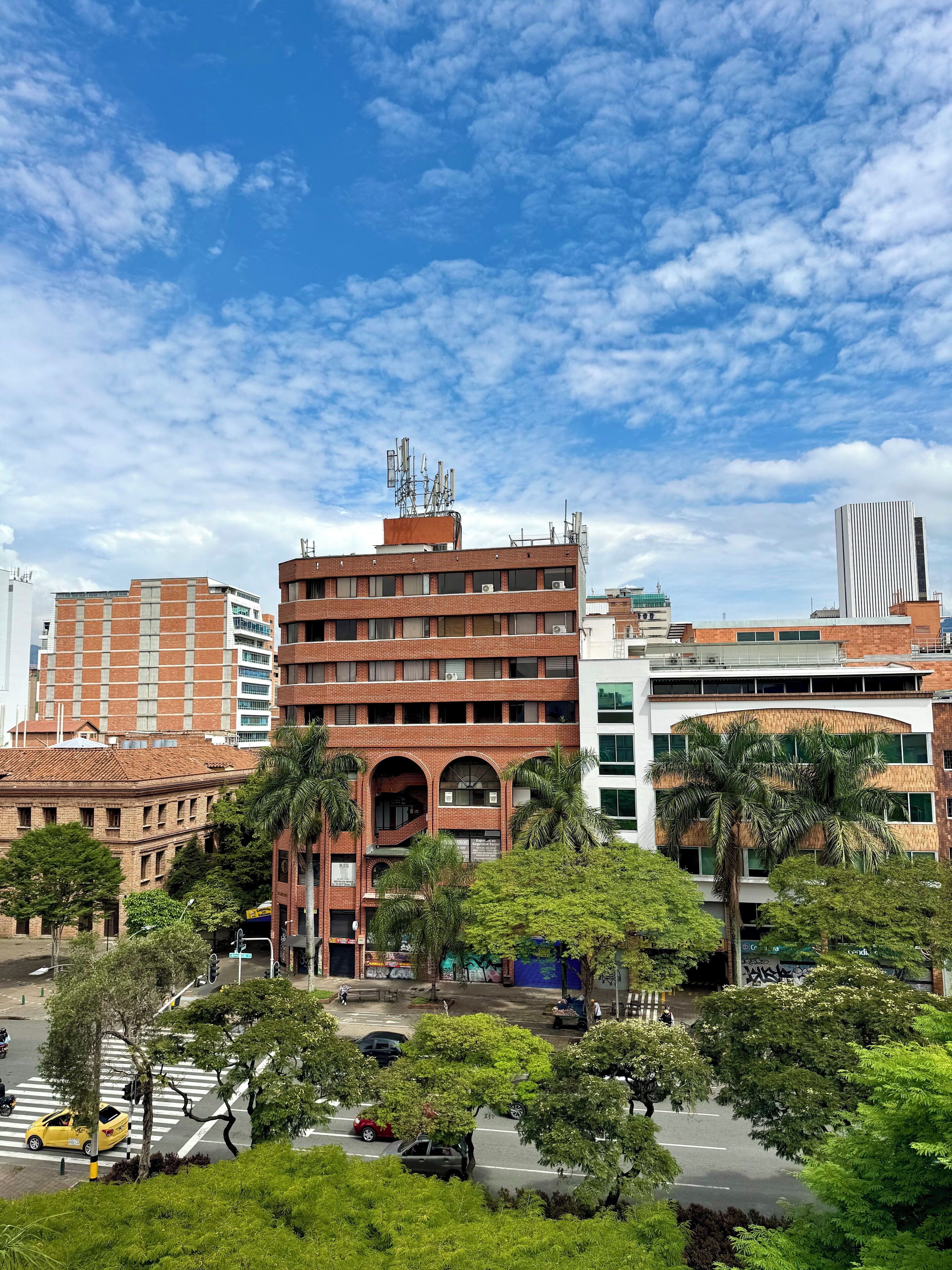 A multi-story building with a brick facade and a flat roof, surrounded by greenery and a clear blue sky.