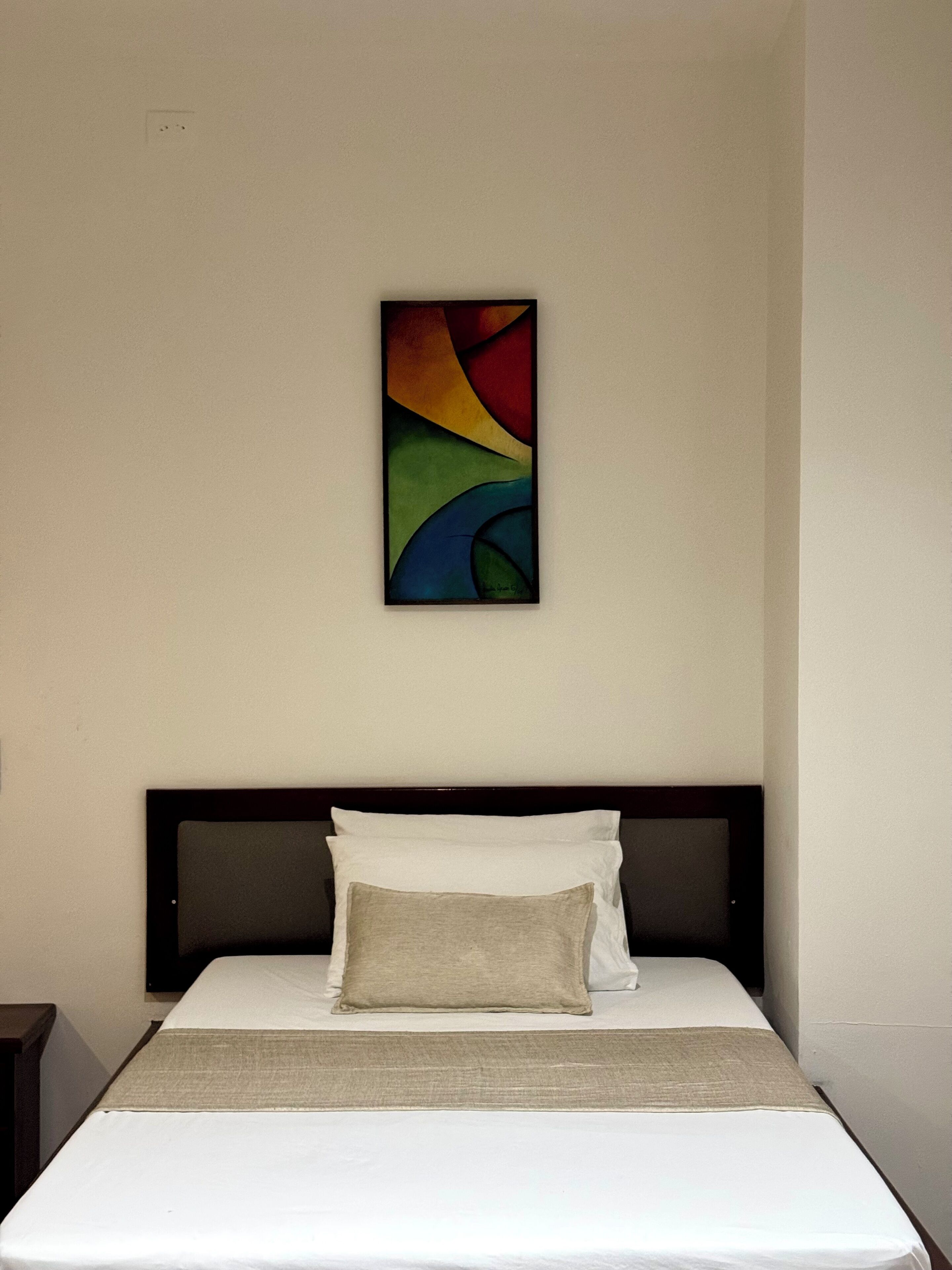 Un lit avec une literie blanche et un oreiller beige, une tête de lit sombre et un tableau abstrait aux couleurs vives accroché au mur.