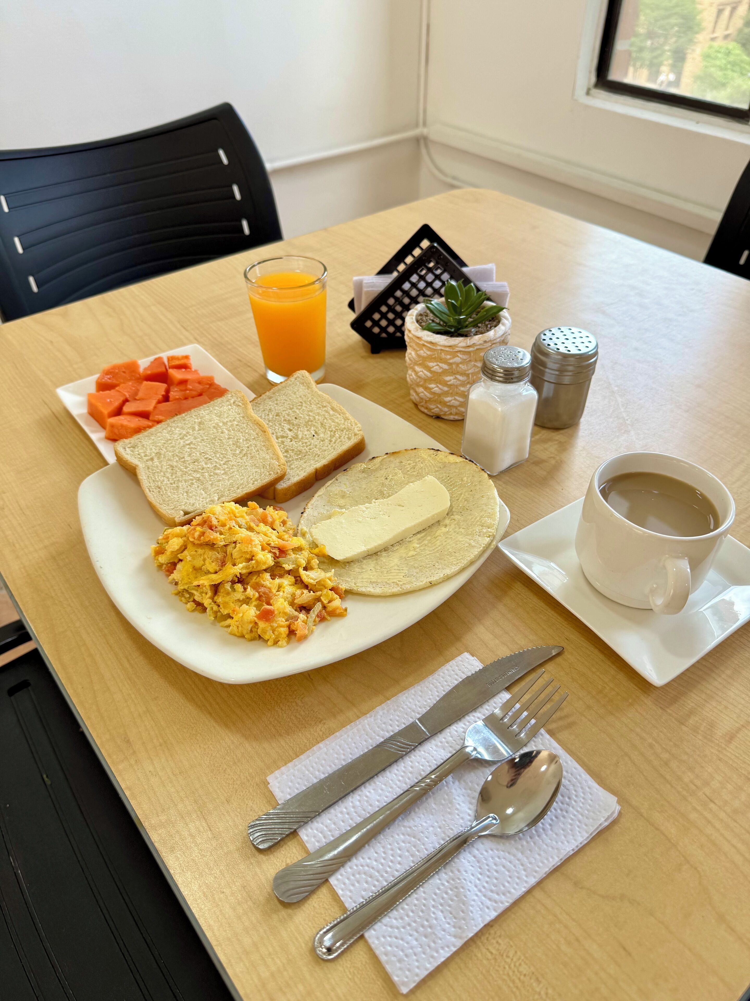 Una mesa de desayuno con tostadas, huevos revueltos, una loncha de queso, un plato de fruta, un vaso de zumo de naranja, una taza de café, saleros y pimenteros, una planta pequeña y cubiertos sobre una servilleta.