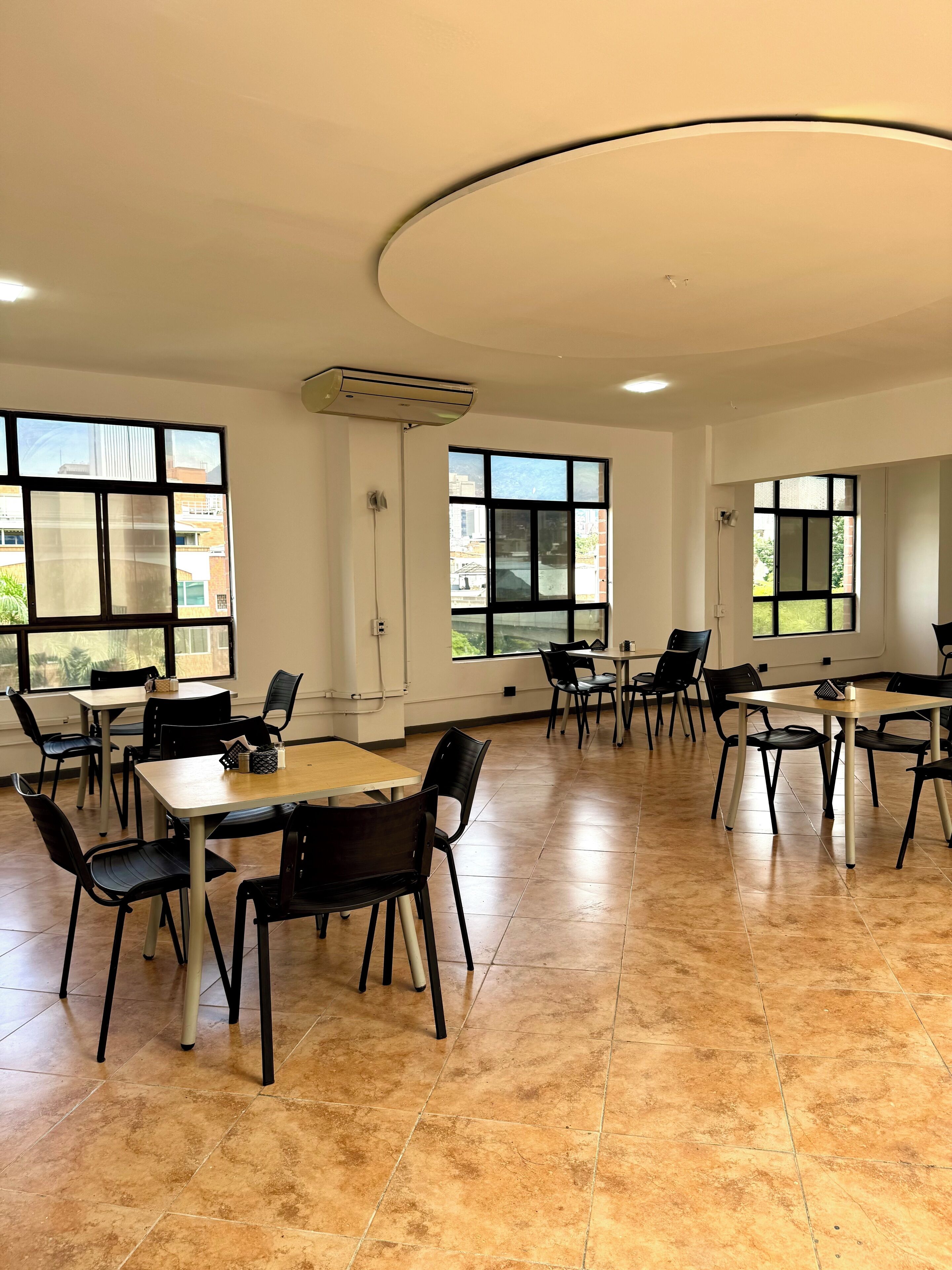 Una habitación amplia con varias mesas y sillas, ventanas grandes que ofrecen vistas a edificios y un techo con diseño circular.