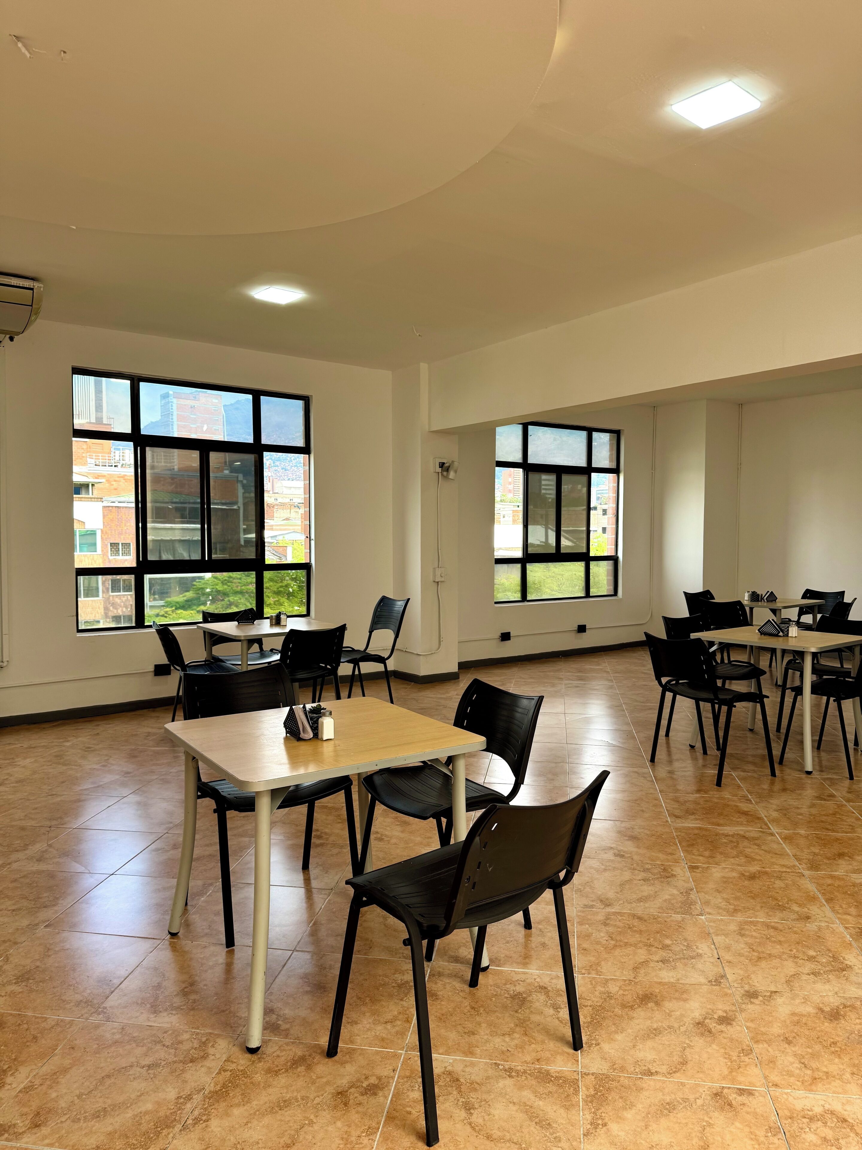 Una habitación amplia con ventanas grandes, pisos de baldosas y varias mesas y sillas.