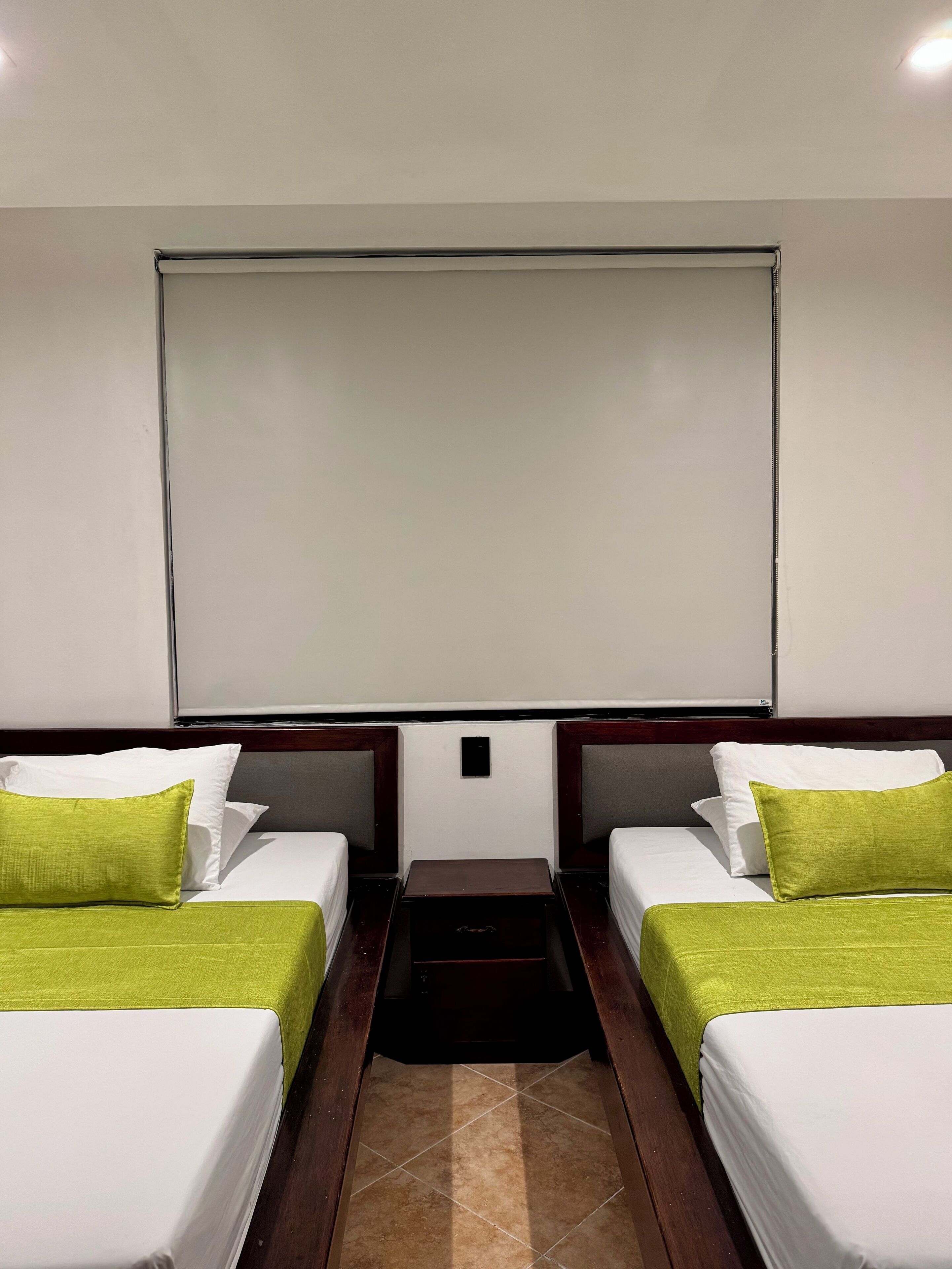 Habitación de hotel con dos camas individuales, cada una con almohada verde y ropa de cama blanca. Hay una pequeña mesa entre las camas y una pantalla de proyector en la pared.
