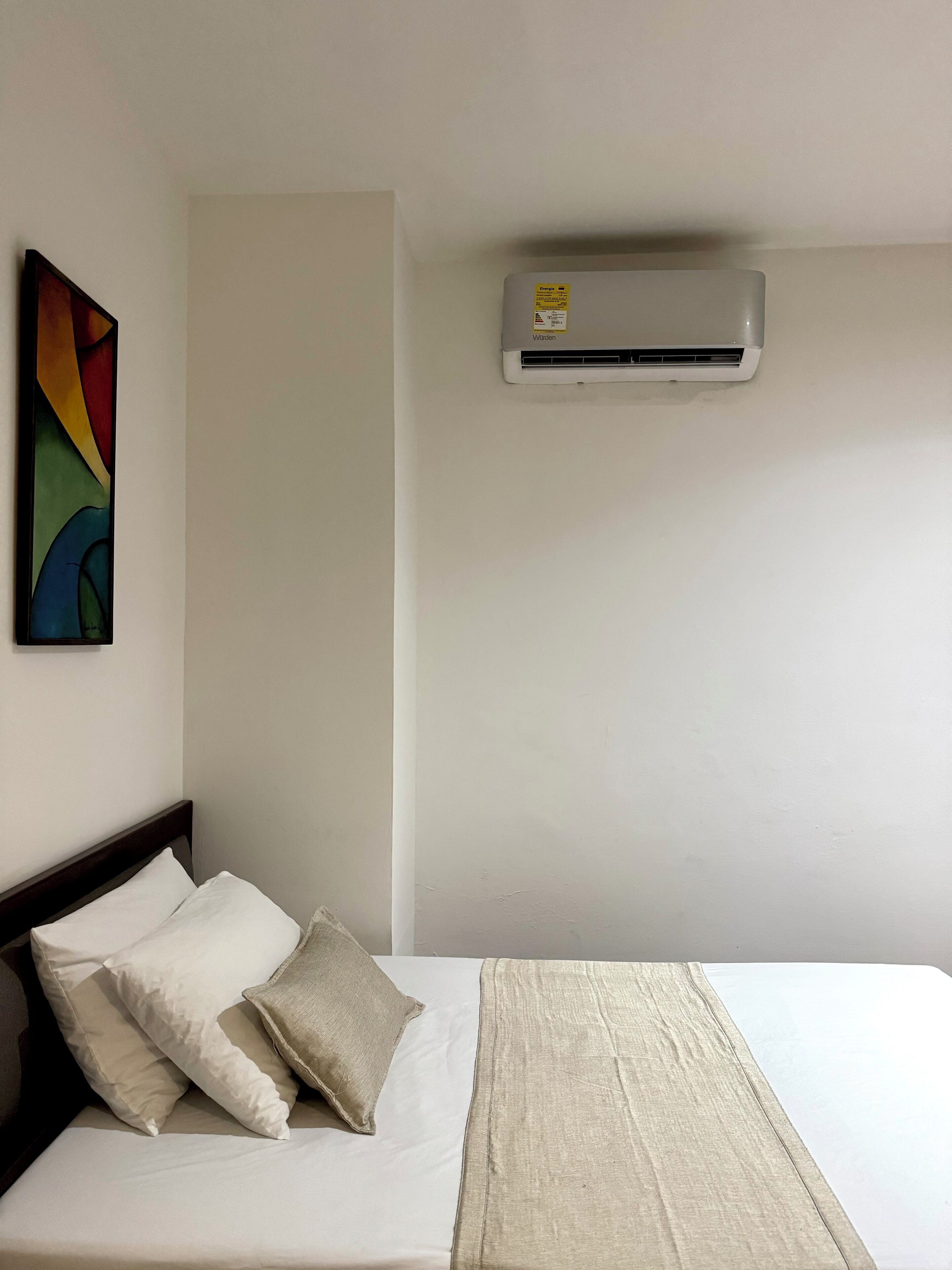 A hotel room with a bed, pillows, a blanket, a wall-mounted air conditioner, and a colorful abstract painting.