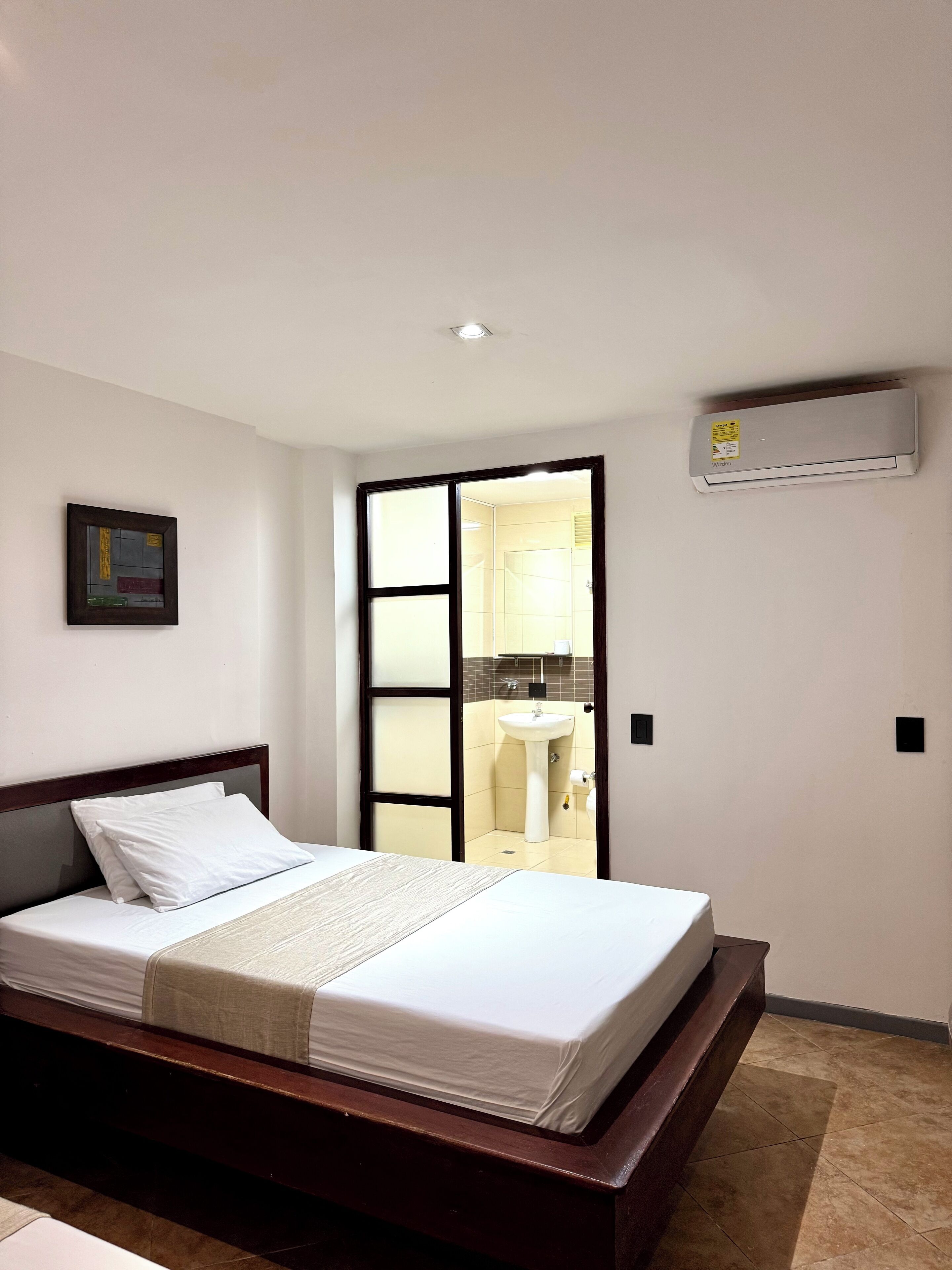 A bedroom with a bed, a bedside table, a wall-mounted air conditioner, and a bathroom visible through a sliding door.
