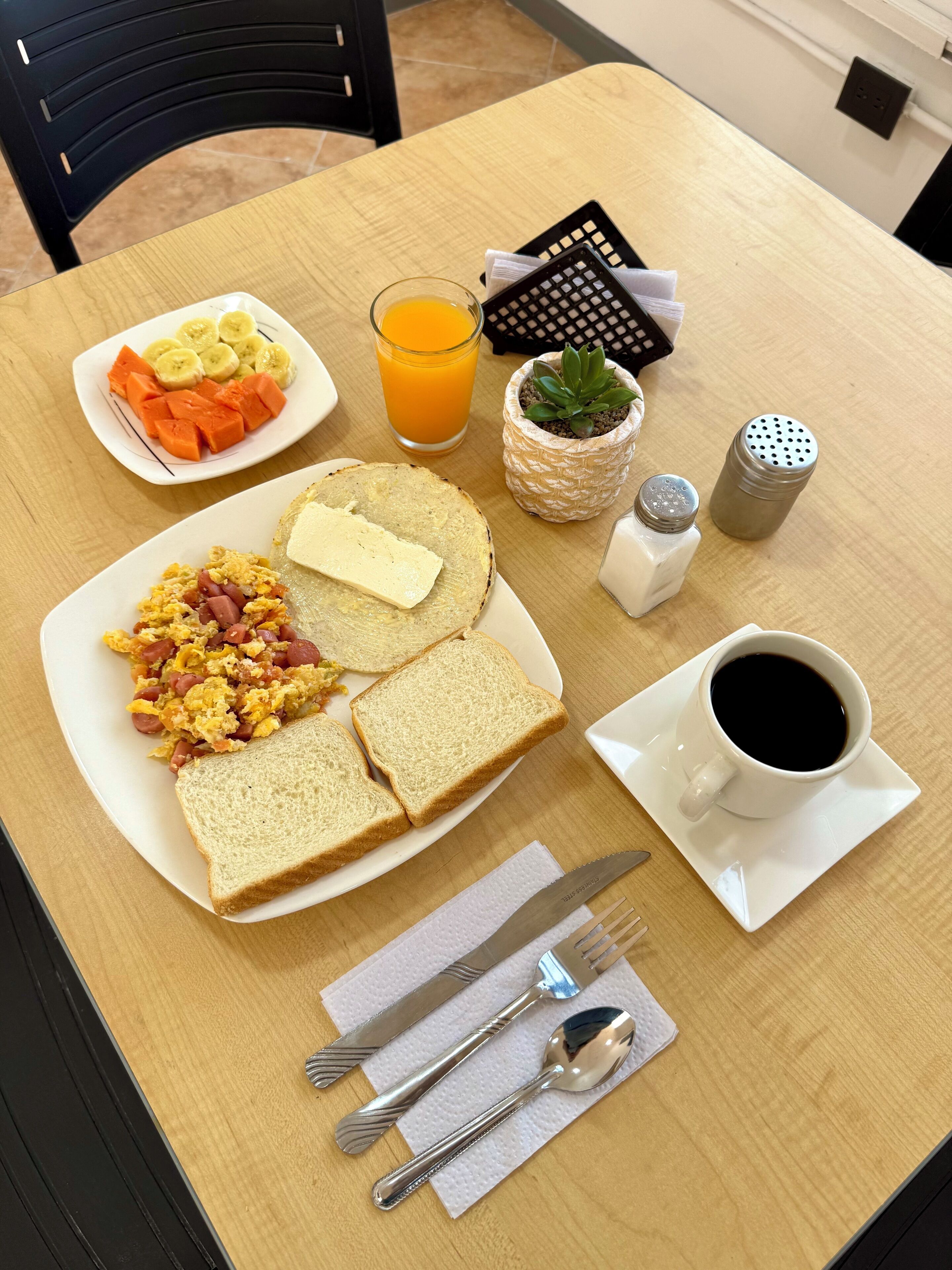 A breakfast table with scrambled eggs, ham, toast, a glass of orange juice, a cup of coffee, and a small plant.