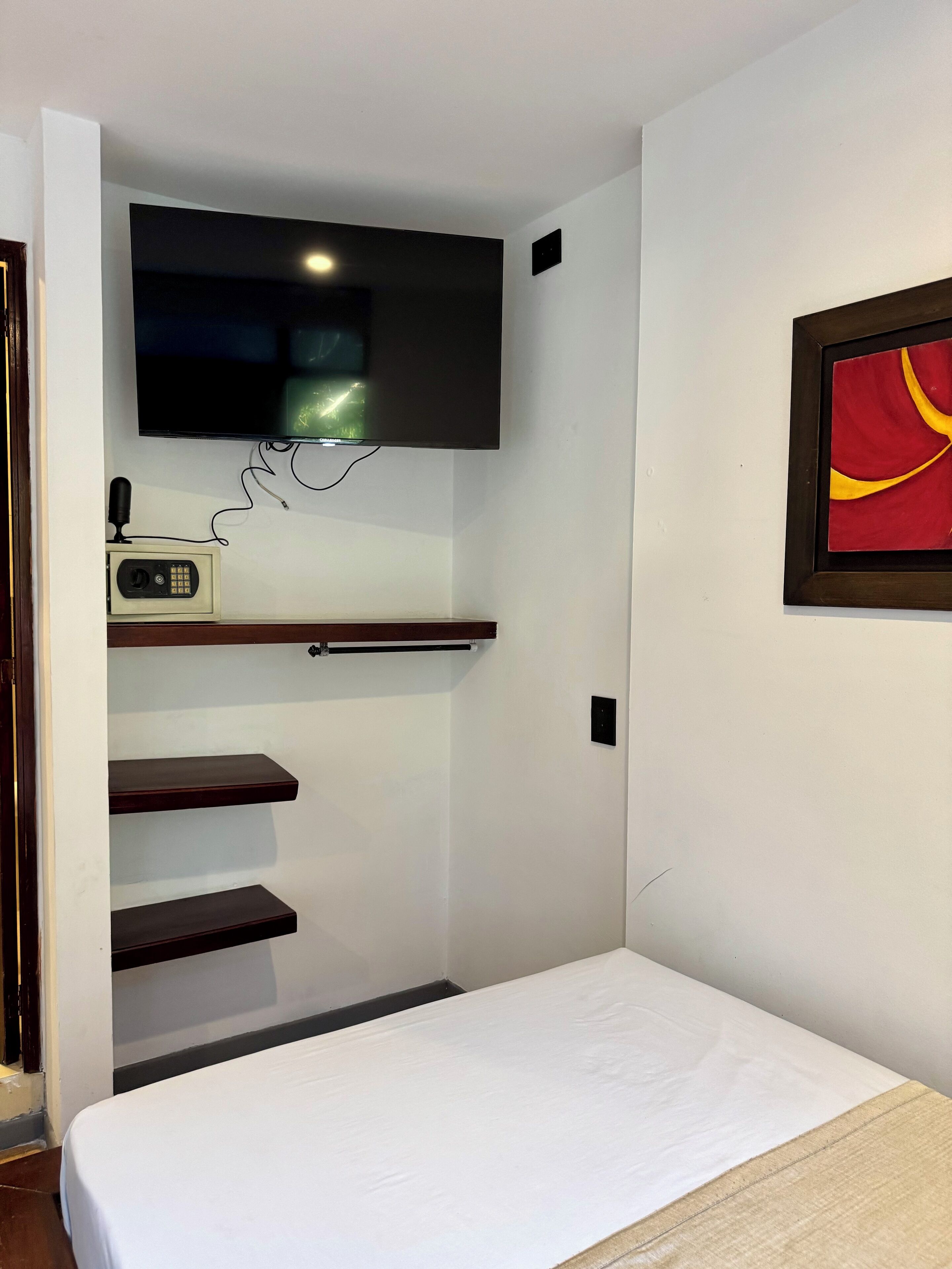 A hotel room with a bed, a mounted television, a shelf with a radio, and a framed picture on the wall.