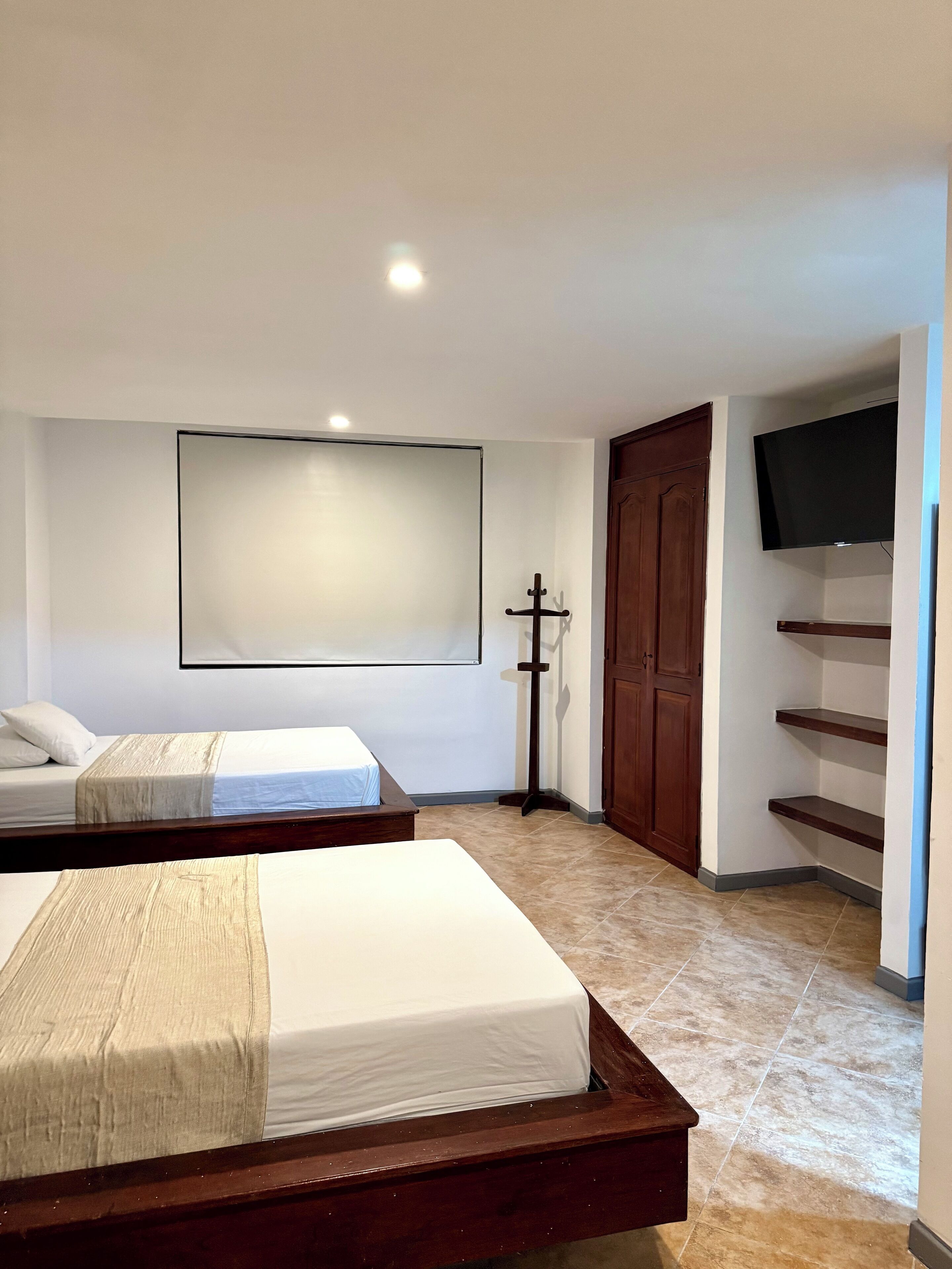 A hotel room with two beds, a TV mounted on the wall, a wooden wardrobe, and a tiled floor.