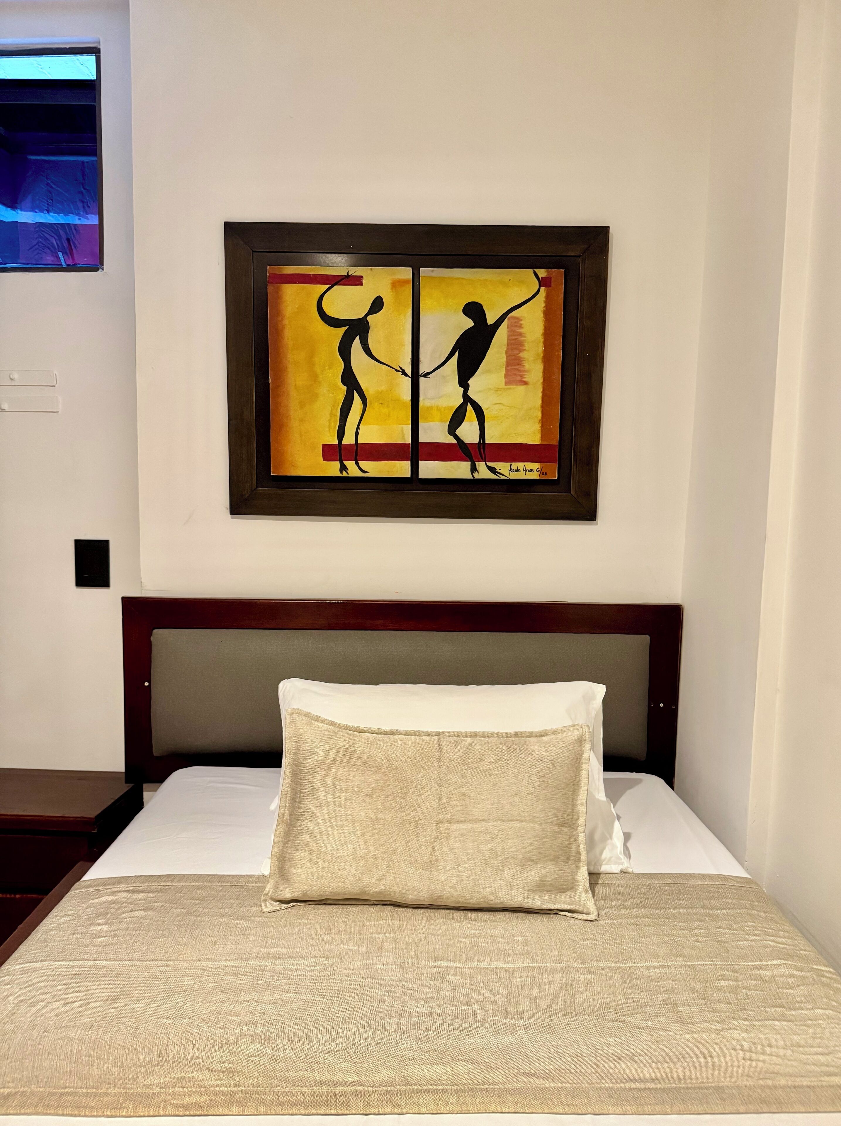 A hotel room with a bed, a headboard, a wall-mounted painting, and a nightstand.