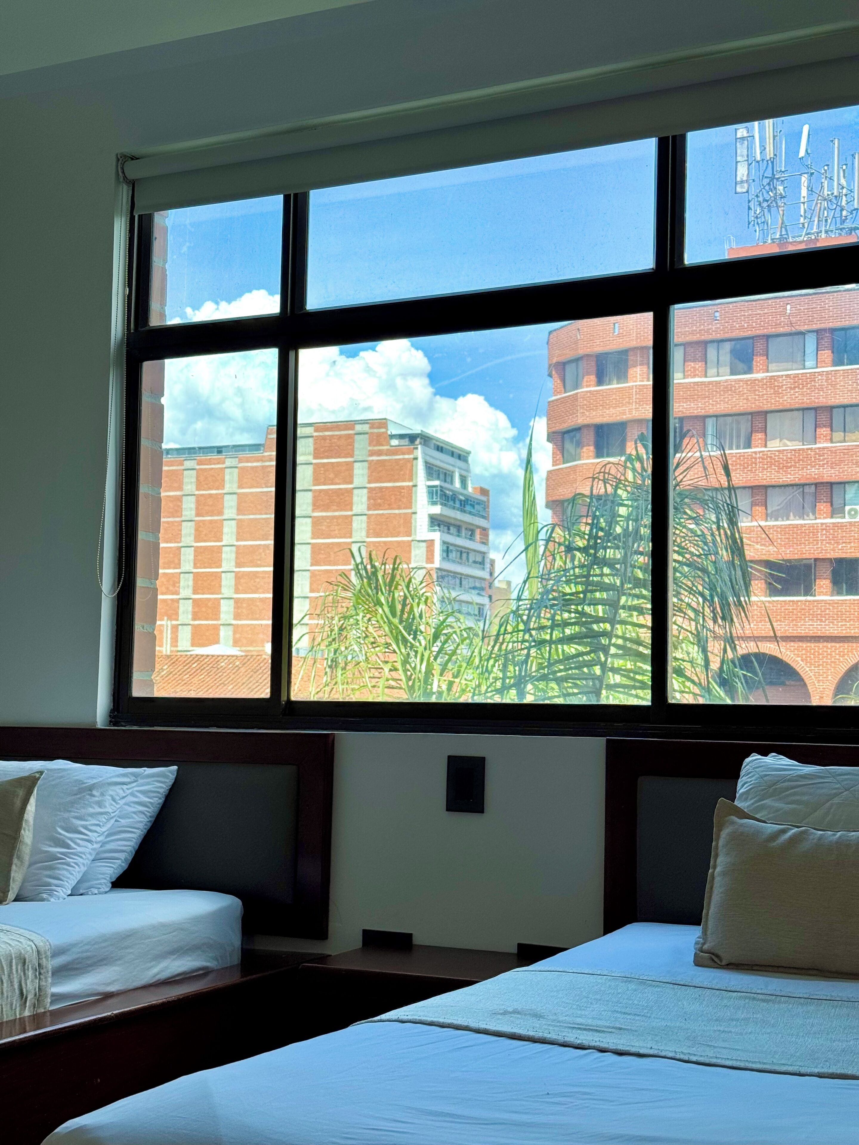 Une chambre d’hôtel avec deux lits et une grande fenêtre donnant sur des immeubles et des plantes.