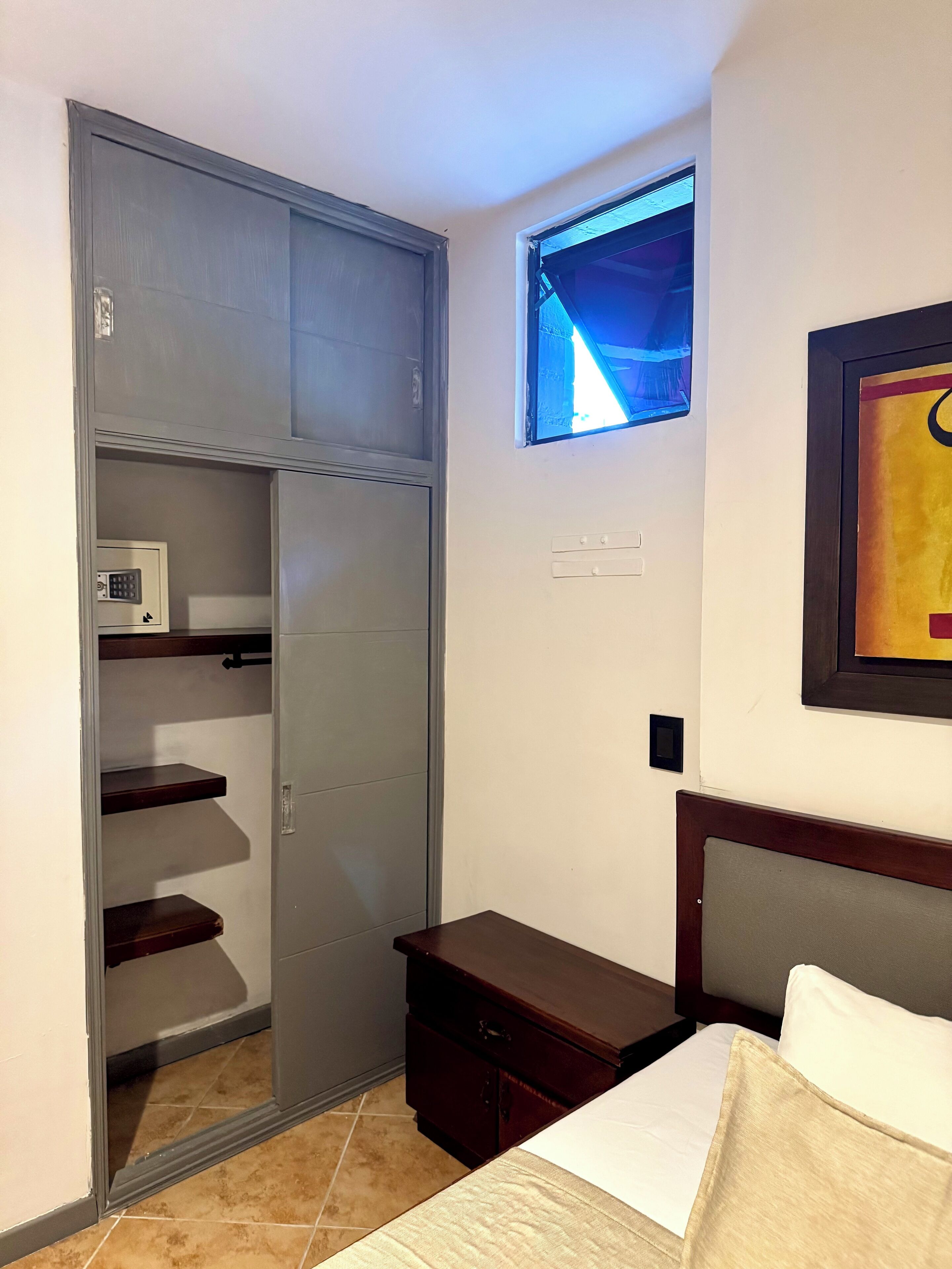 A bedroom with a bed, a nightstand, a wooden dresser, a closet with shelves, a window, and a wall-mounted light switch.