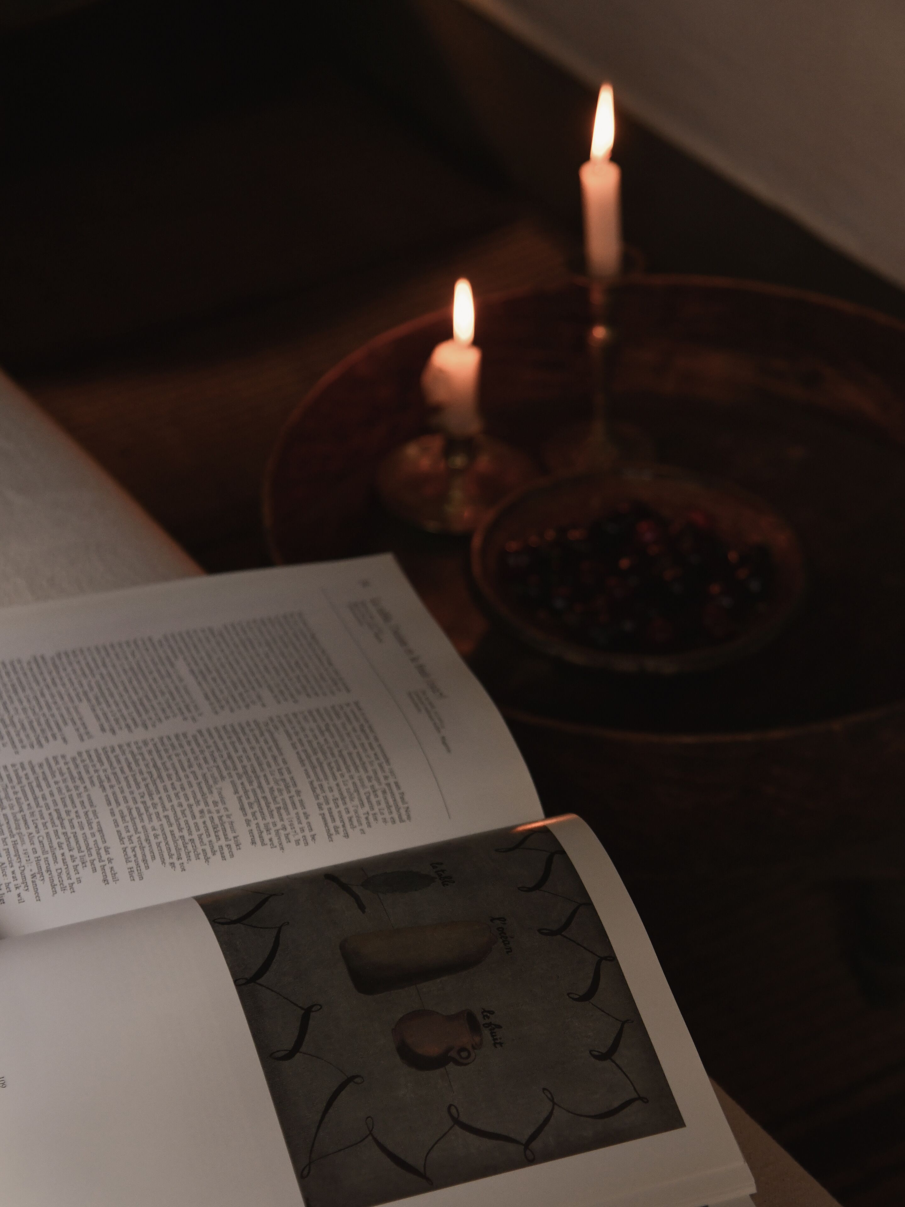 An open book with text on a table, a lit candle, and a decorative plate.