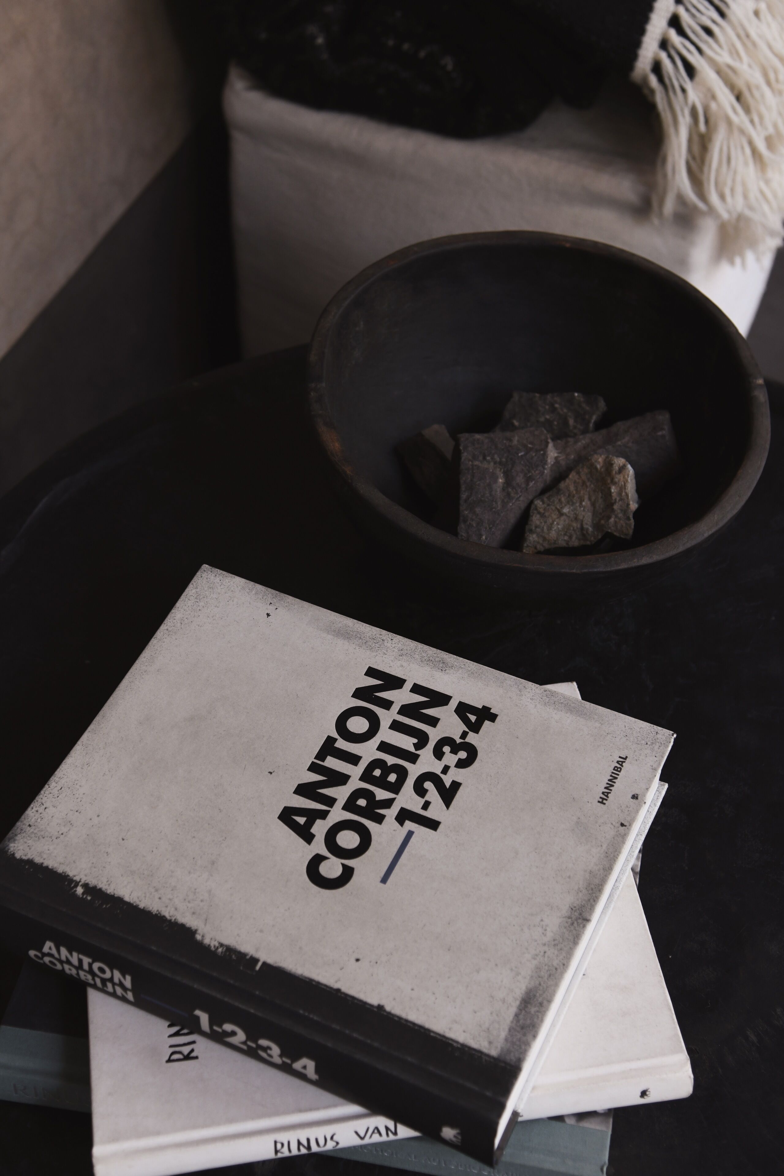 A book titled 'Anton Corbijn 1-2-3-4' on a table with a bowl of rocks and a white bag in the background.