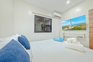 A hotel room with a bed, a window, an air conditioning unit, a painting of a beach, and a wooden door. - New Sea La Vie Suite # 2 |2BR|1BR W. Pool&Jacuzzi (Noord)