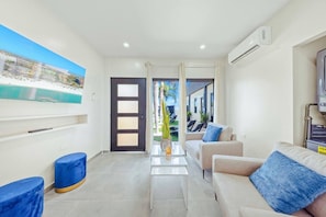 A modern hotel room with a large window, a flat-screen TV, a sofa, a coffee table, and a small blue ottoman. - New Sea La Vie Suite # 2 |2BR|1BR W. Pool&Jacuzzi (Noord)