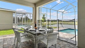 A covered patio with a glass enclosure, a dining table set for four, wicker chairs, and a view of a swimming pool and buildings in the background. - 9BR Lakeview Villa - Pool, Jacuzzi & Pool Table! (Kissimmee)