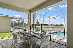 Un patio cubierto con cerramiento de vidrio, una mesa de comedor preparada para cuatro personas, sillas de mimbre y vistas a una piscina y edificios al fondo.